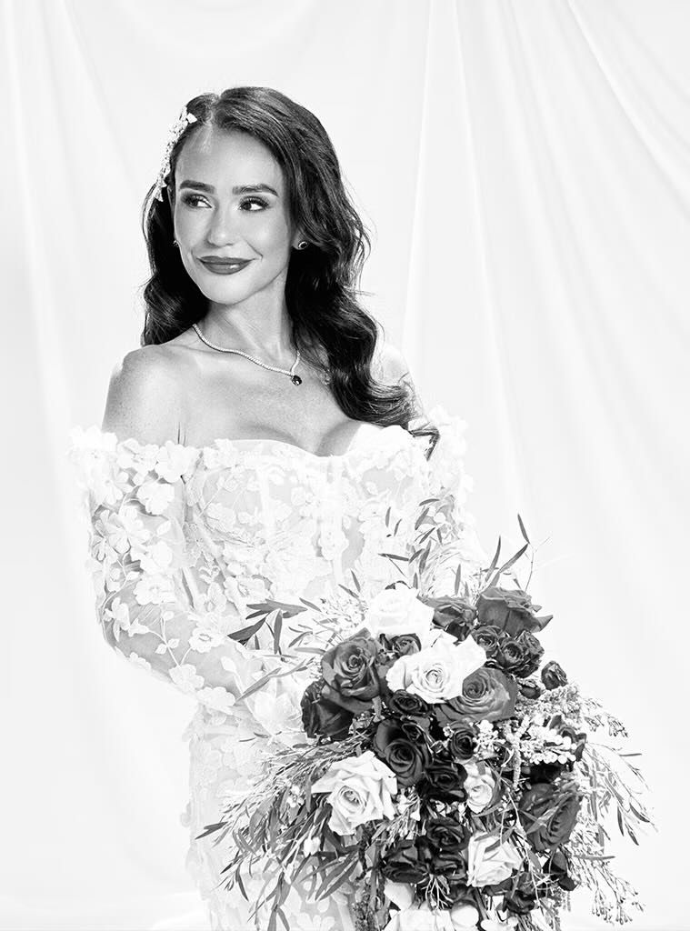 Black and white bridal portrait of a woman in an off-shoulder lace gown holding a bouquet of roses.