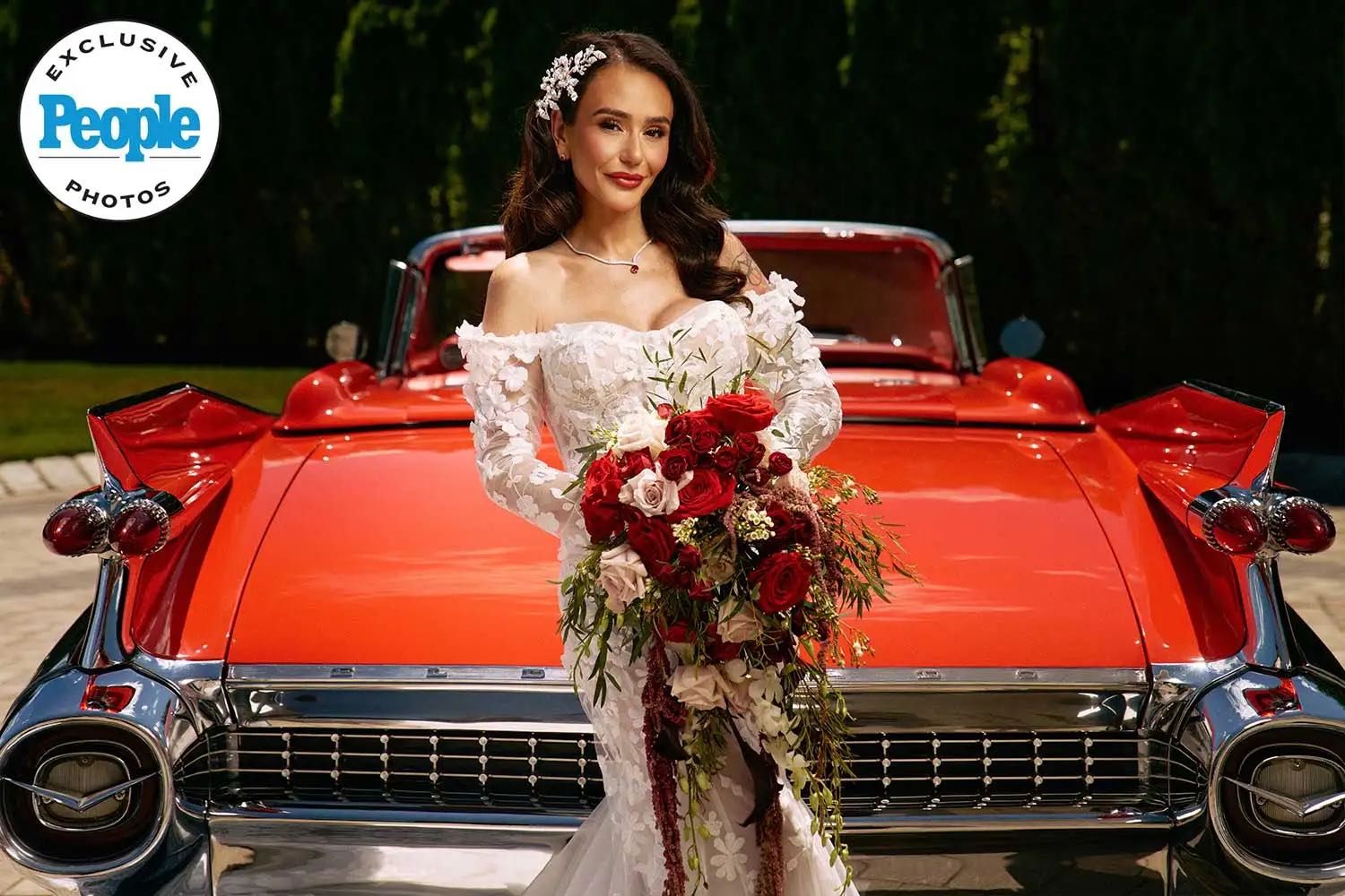 Bride in white off-shoulder gown holds red bouquet in front of classic red Cadillac.