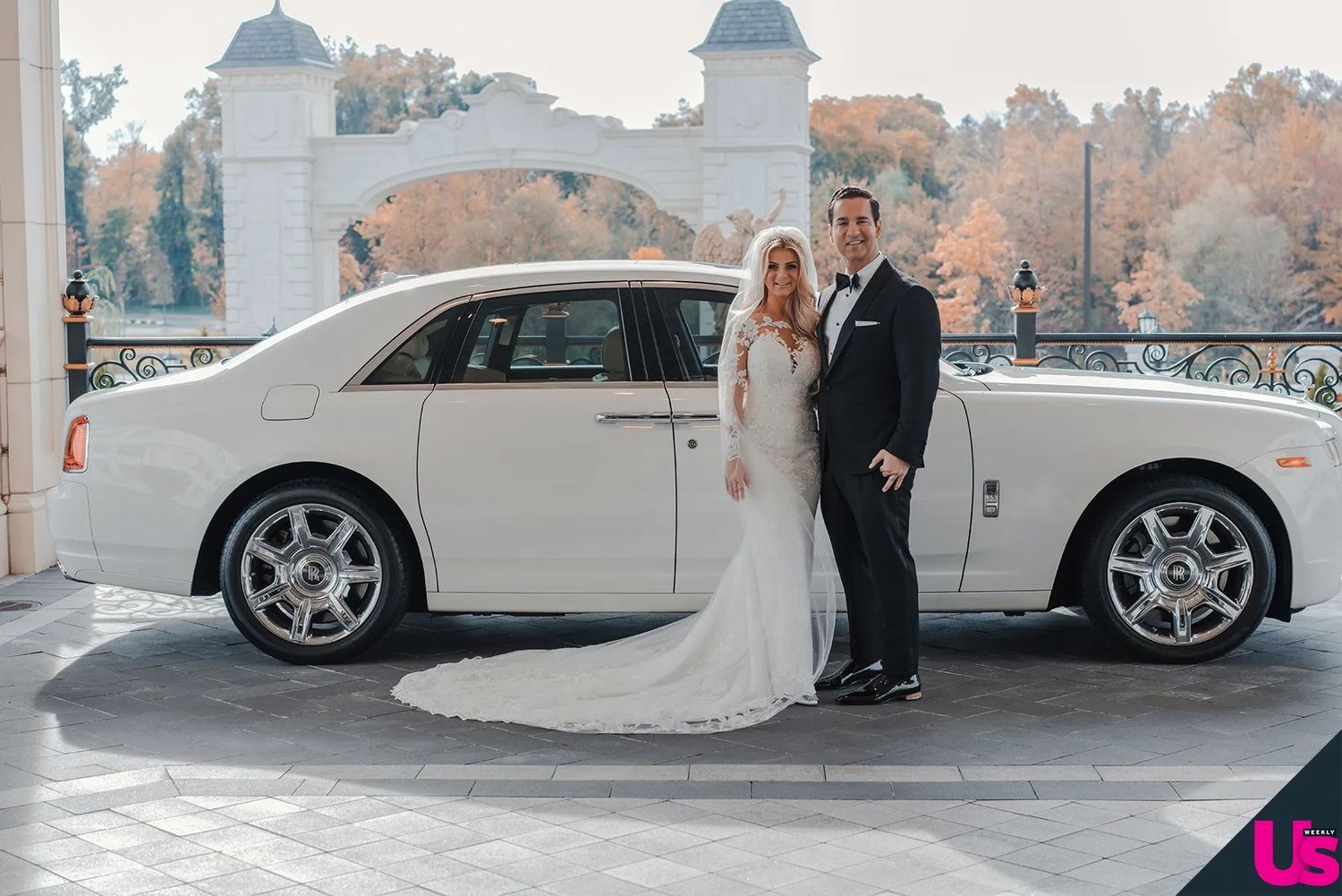 Bride and groom pose in wedding attire beside a white Rolls-Royce Ghost at an elegant outdoor venue.