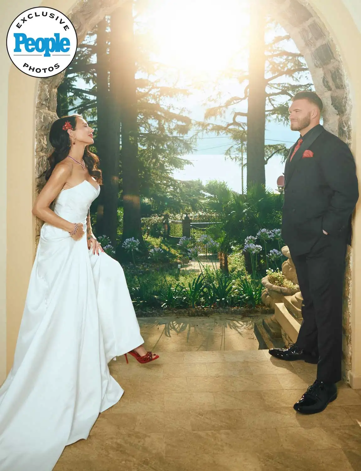 Bride in white gown with red heels faces groom in black suit in sunlit garden archway, People magazine exclusive wedding photo.