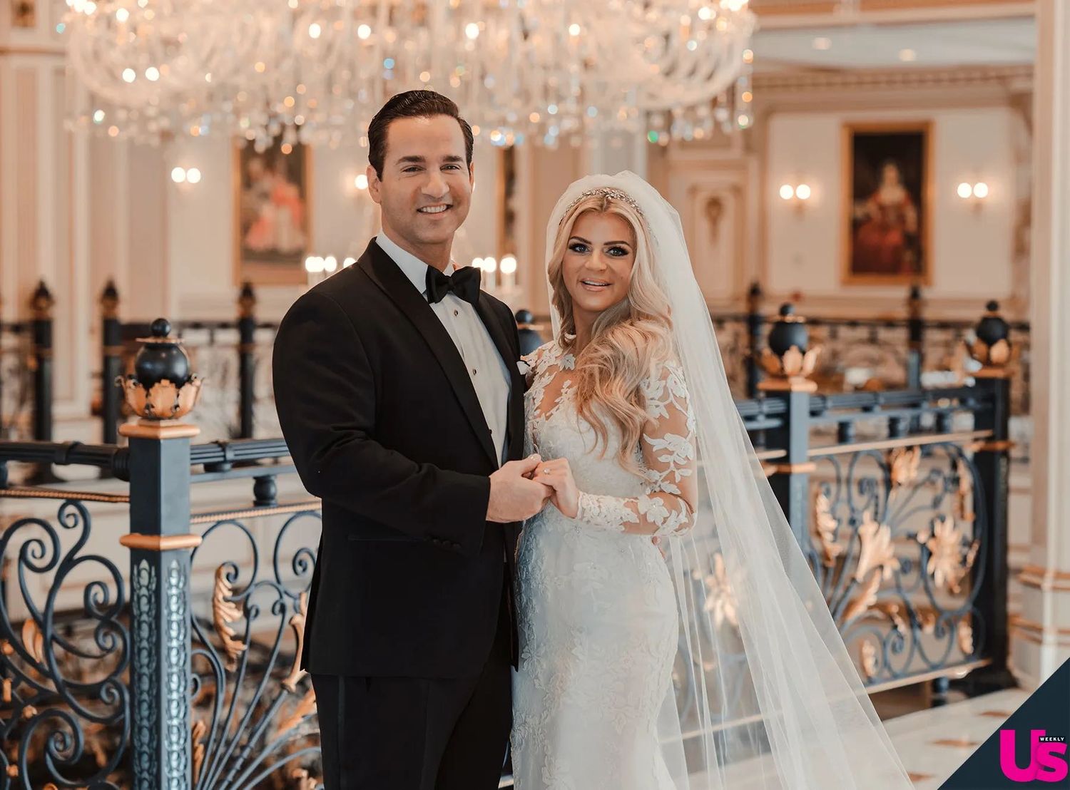 Elegant bride and groom pose together in a grand ballroom on their wedding day, wearing a tuxedo and lace gown.