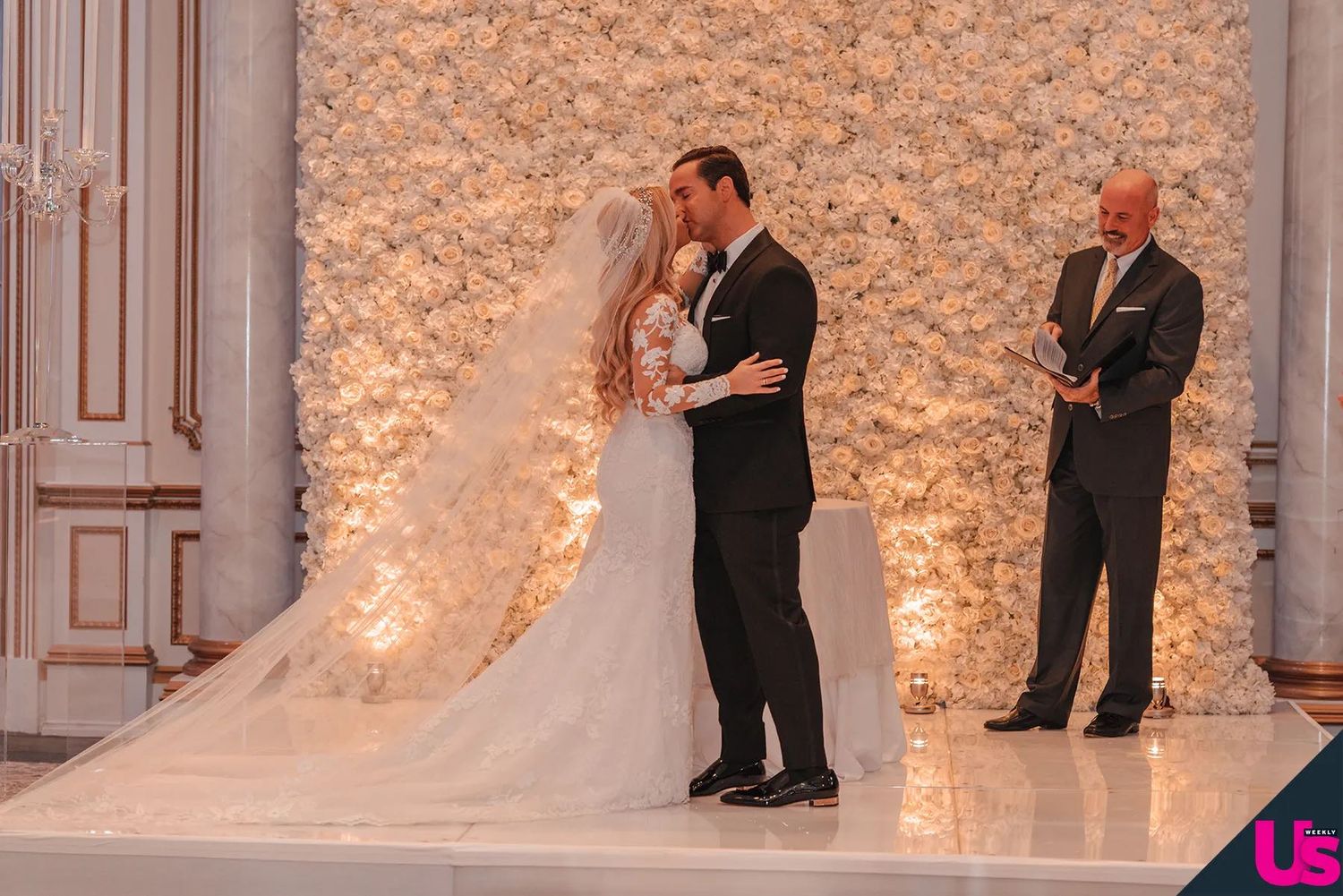 Bride in white gown and groom in tuxedo exchange vows before officiant at elegant floral wedding ceremony.