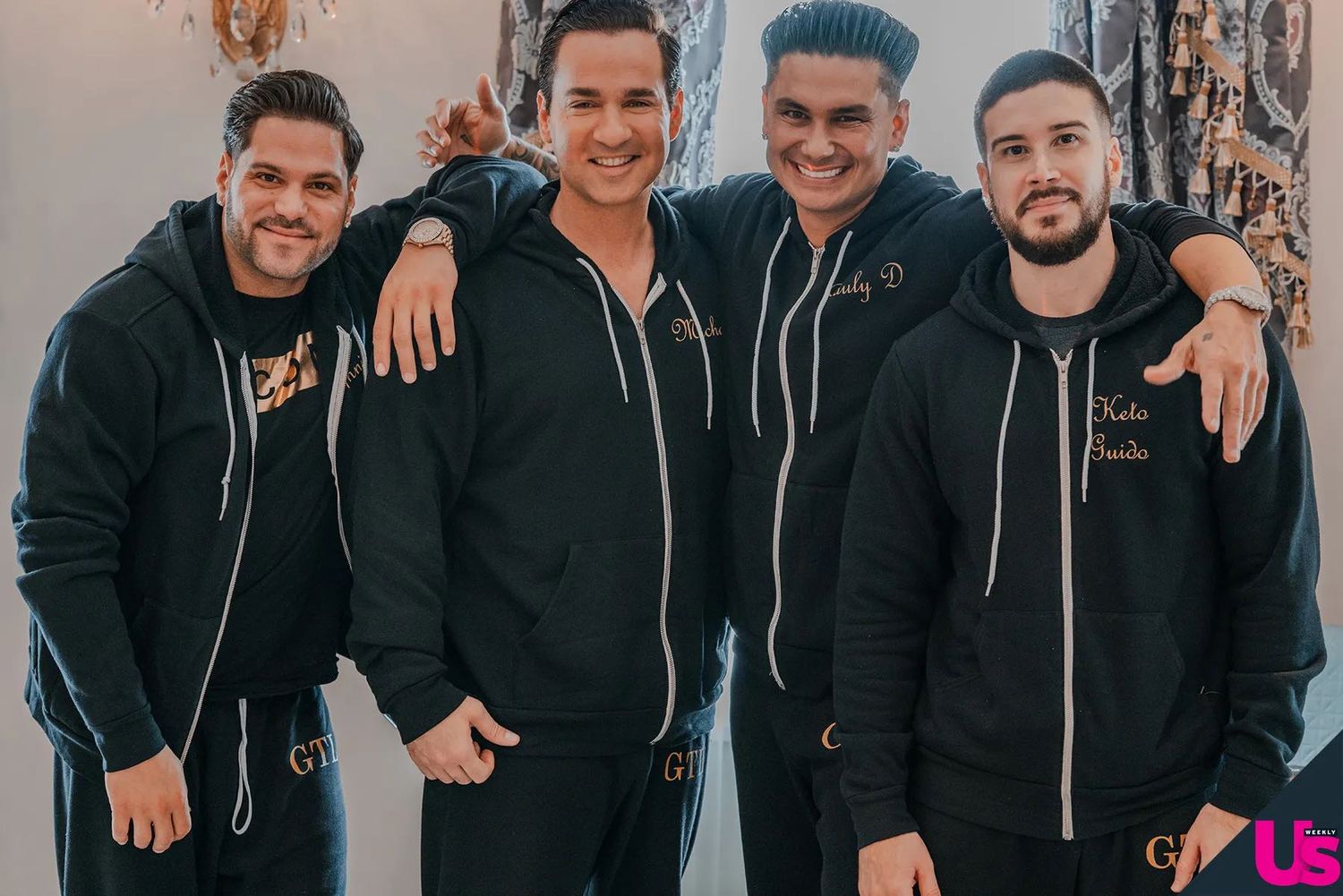 Four men in matching black zip-up hoodies smile together for a group photo at a formal event.