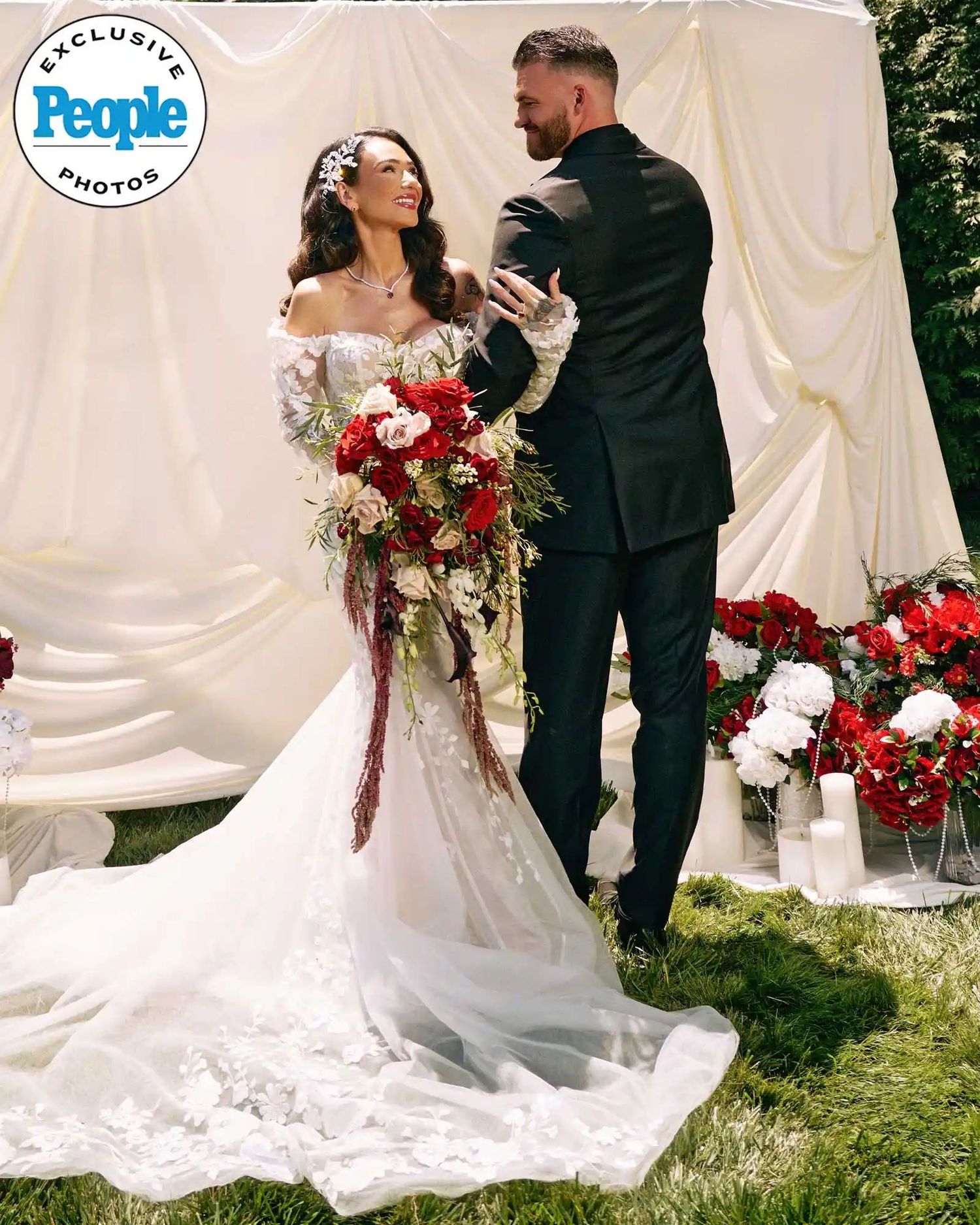 Bride in white gown holds red bouquet as groom reads vows at outdoor wedding ceremony with draped backdrop.
