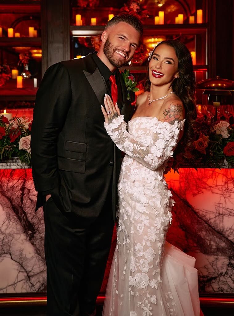 Couple posing in wedding attire, man in black tuxedo and woman in white floral gown, in a romantic red-lit venue.
