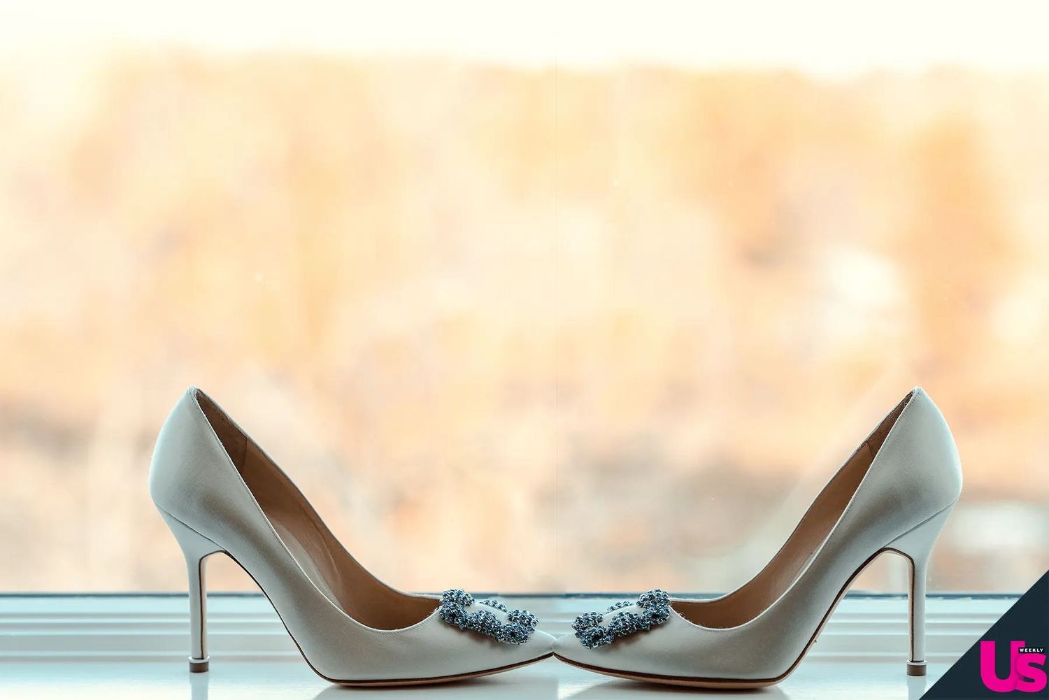 White bridal heels with decorative blue crystal buckles displayed on a windowsill with soft bokeh background.