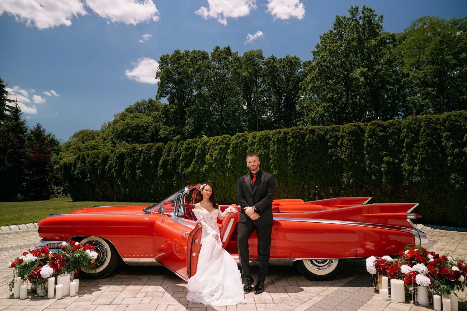 Bride and groom posing beside a classic red vintage Cadillac convertible decorated with flowers on a sunny wedding day.