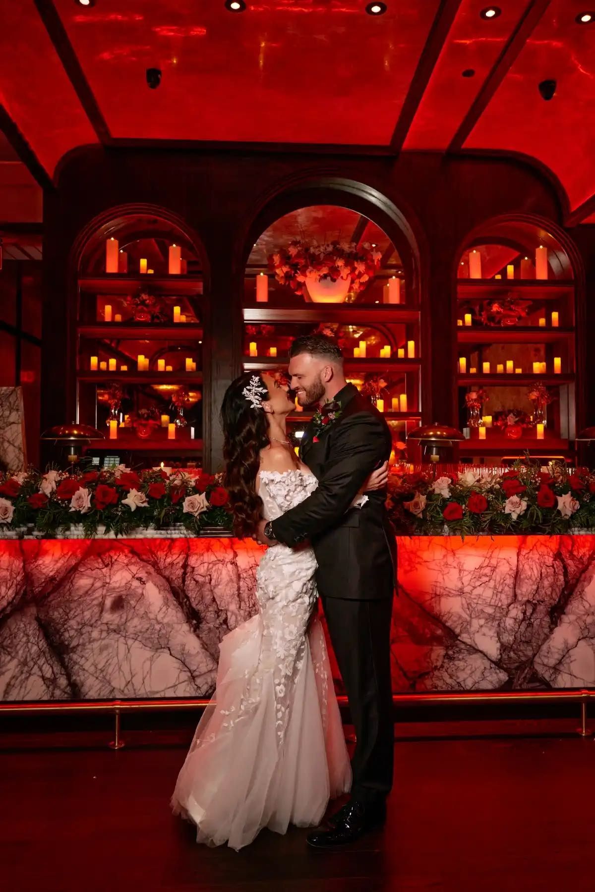 Bride and groom embrace in front of a glowing red candlelit bar with marble countertop and floral arrangements.