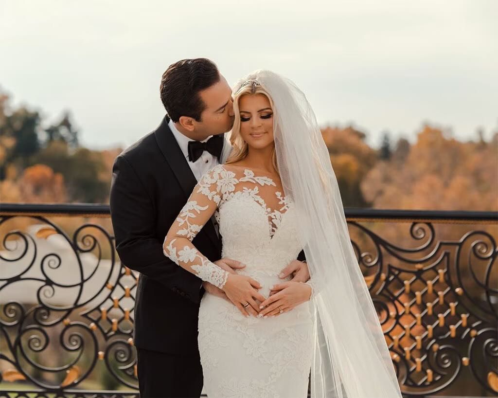 Bride in lace gown with veil and groom in tuxedo share romantic kiss on ornate balcony with autumn foliage backdrop.