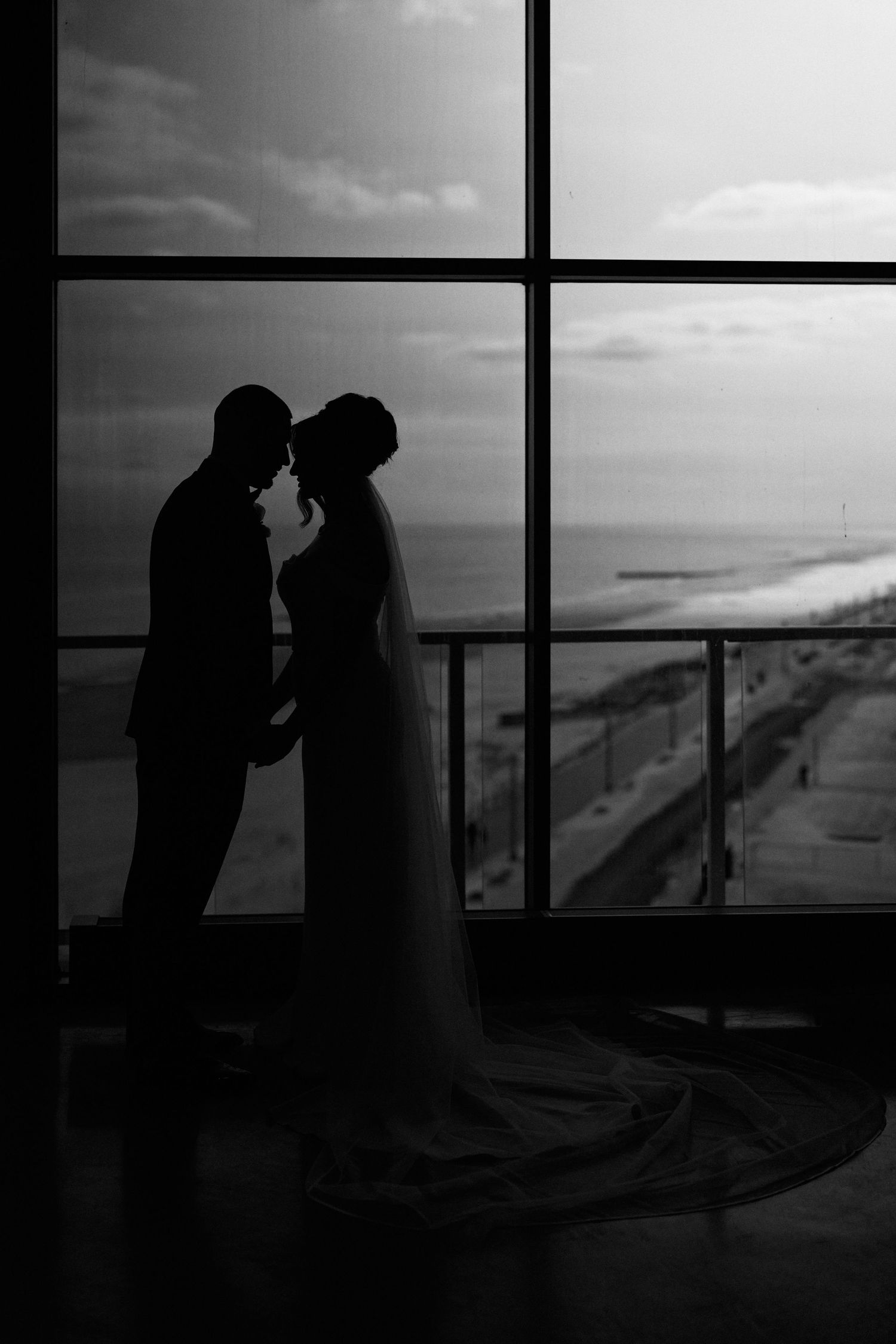 Black and white silhouette of bride and groom standing before large window overlooking ocean on wedding day.