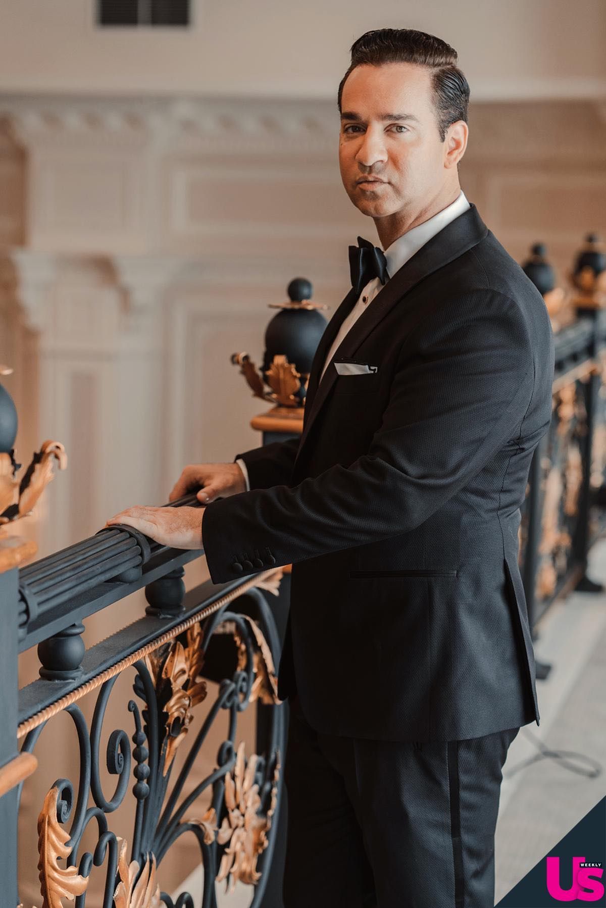 An elegant man in a black tuxedo stands at an ornate iron railing in a grand interior setting.