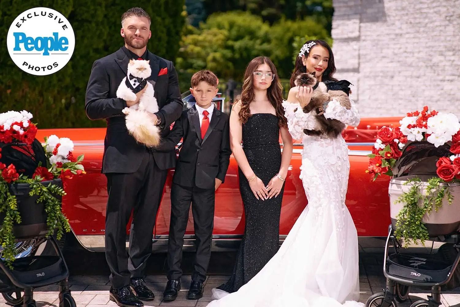 Wedding party poses with dogs by red car, bride in white gown, groom in black suit, two children dressed formally.