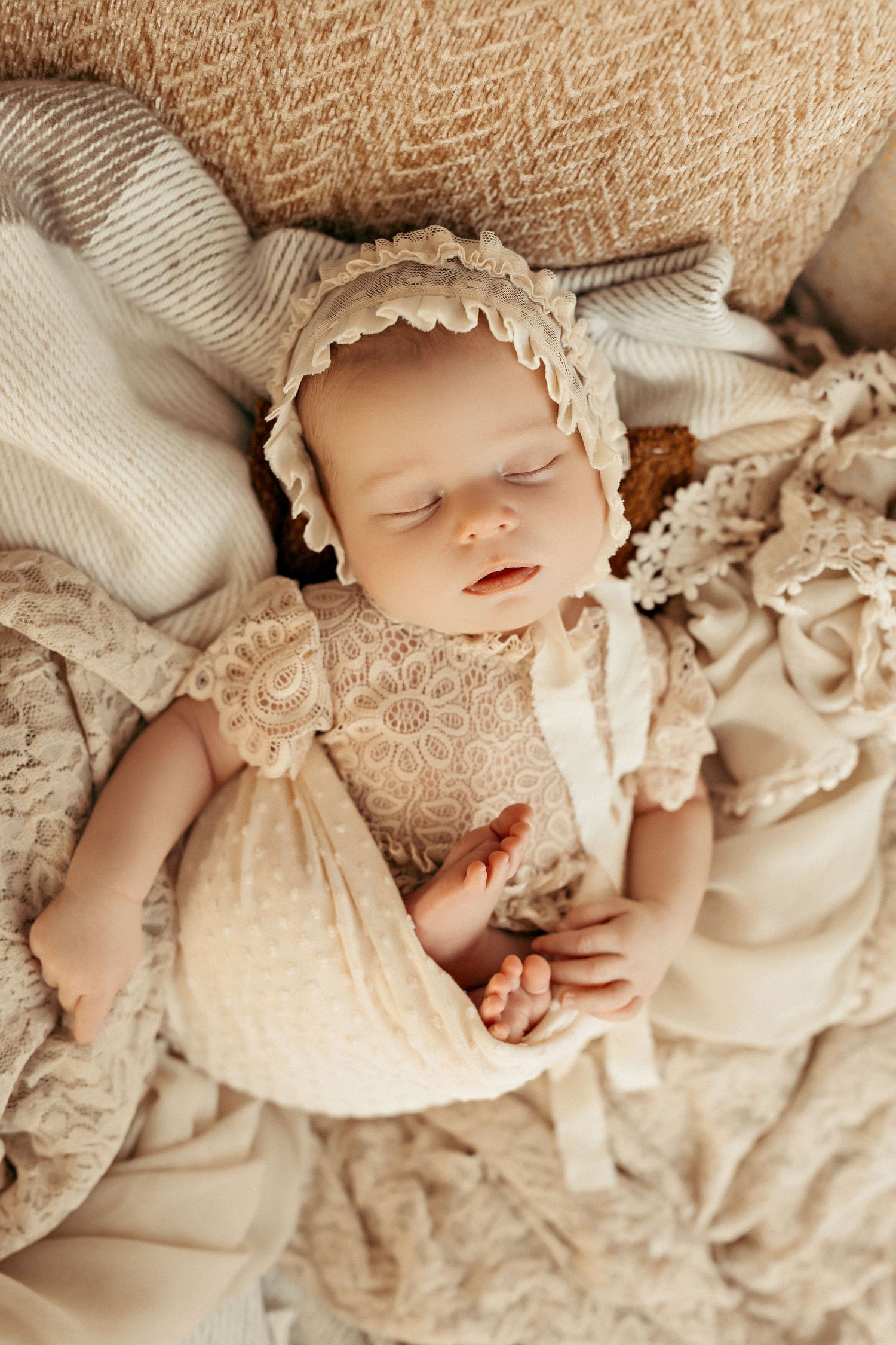 Sleeping newborn wearing vintage lace bonnet and dress on cream textured blankets.