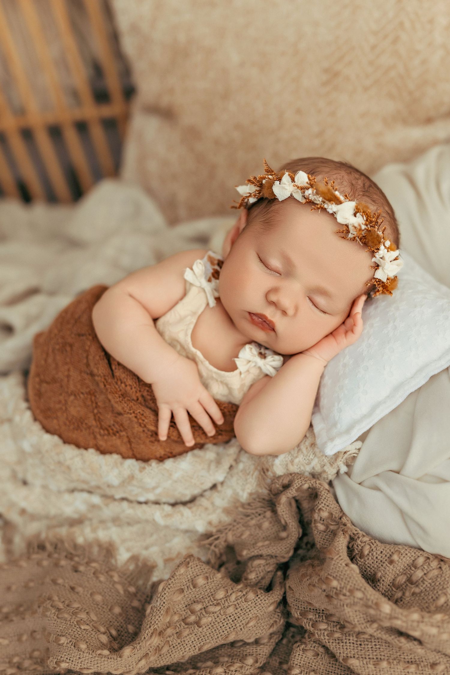 Sleeping newborn wearing floral crown on textured brown fabric.