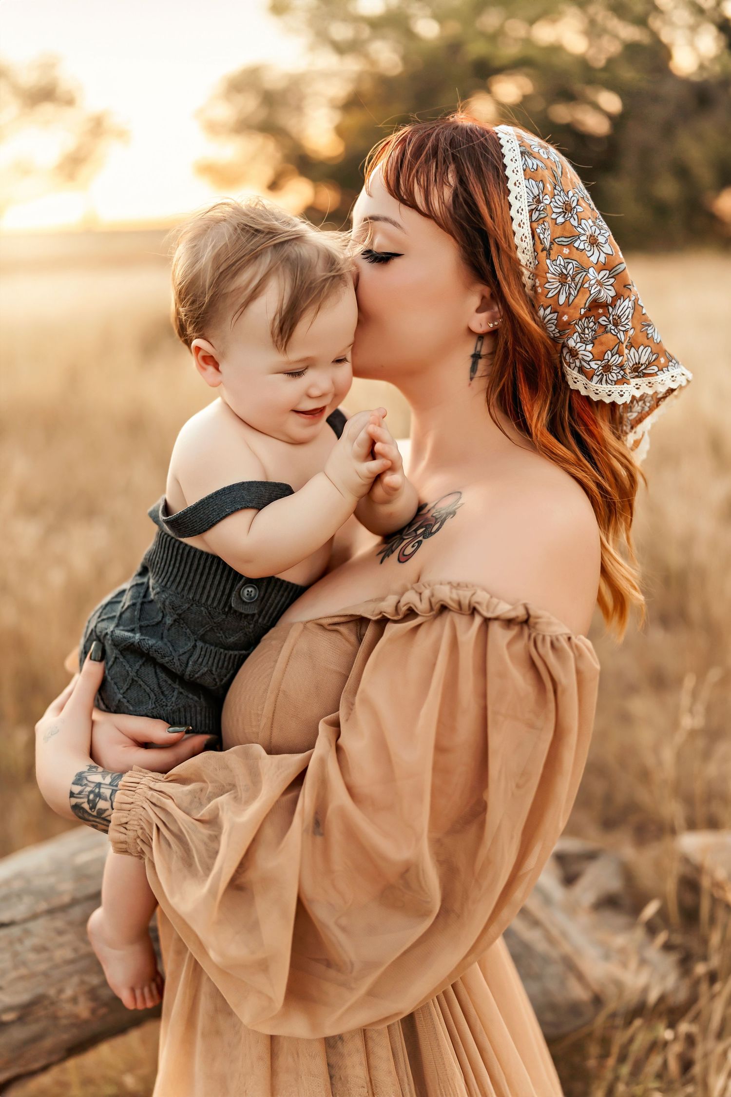 Mother in floral headscarf and tan dress kisses baby in overalls during sunset portrait.
