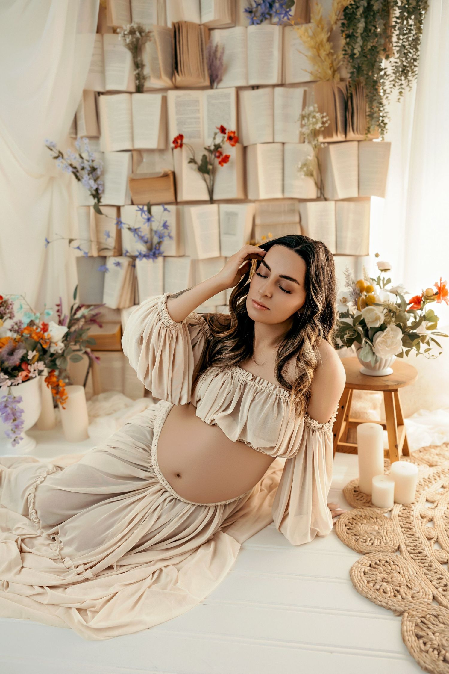 Maternity photo in soft beige dress with open books and floral decorations on wall creating dreamy boho atmosphere.