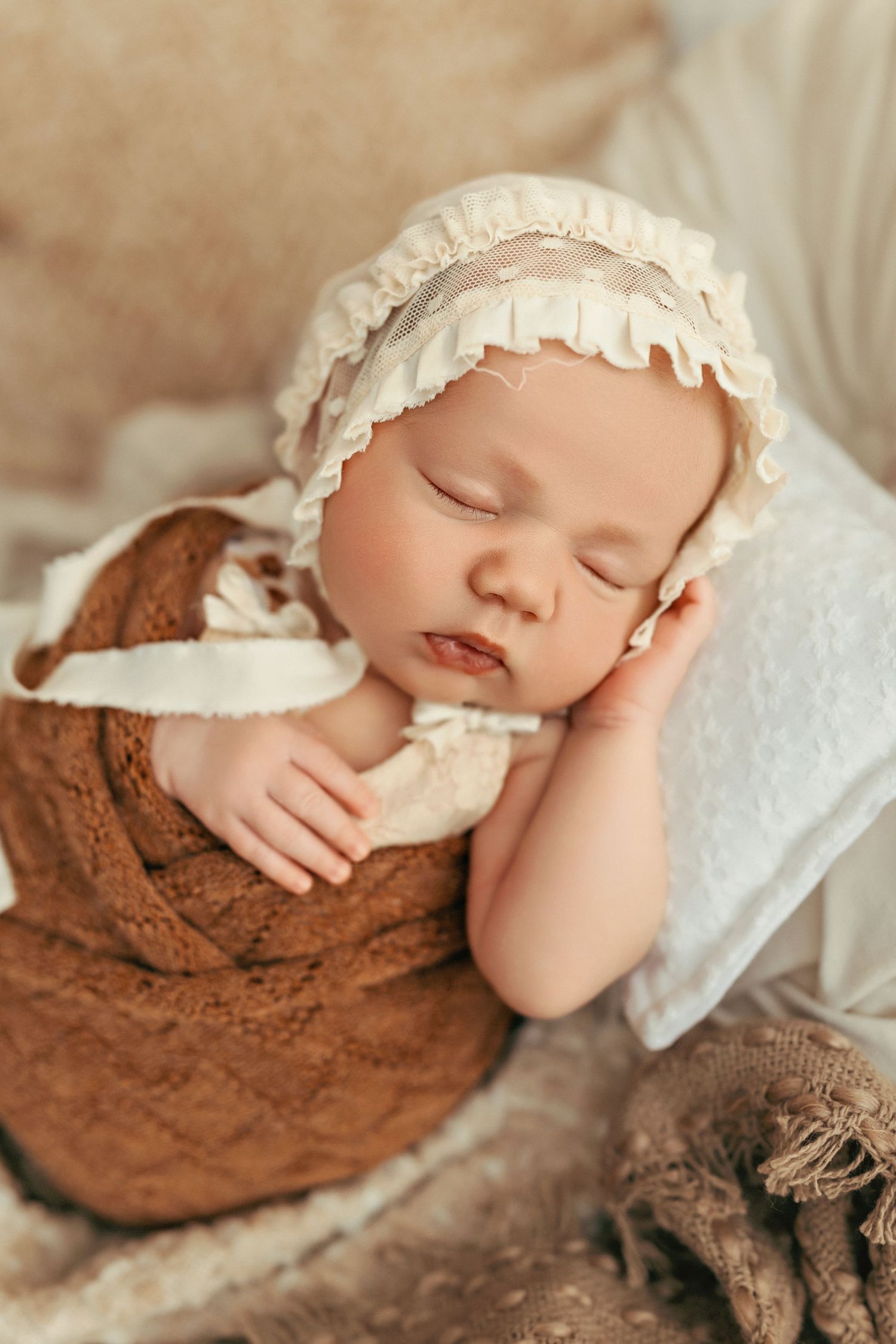 Sleeping baby in vintage-style bonnet and brown dress during professional portrait session.