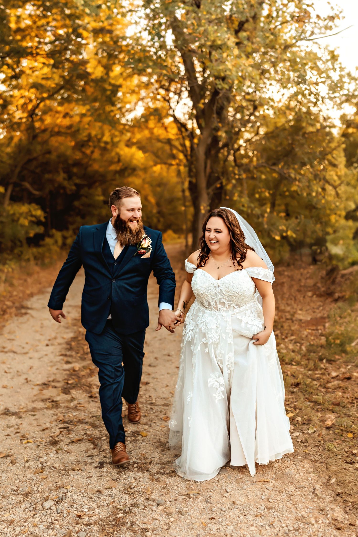 Newlyweds walking hand in hand down forest path in autumn foliage.