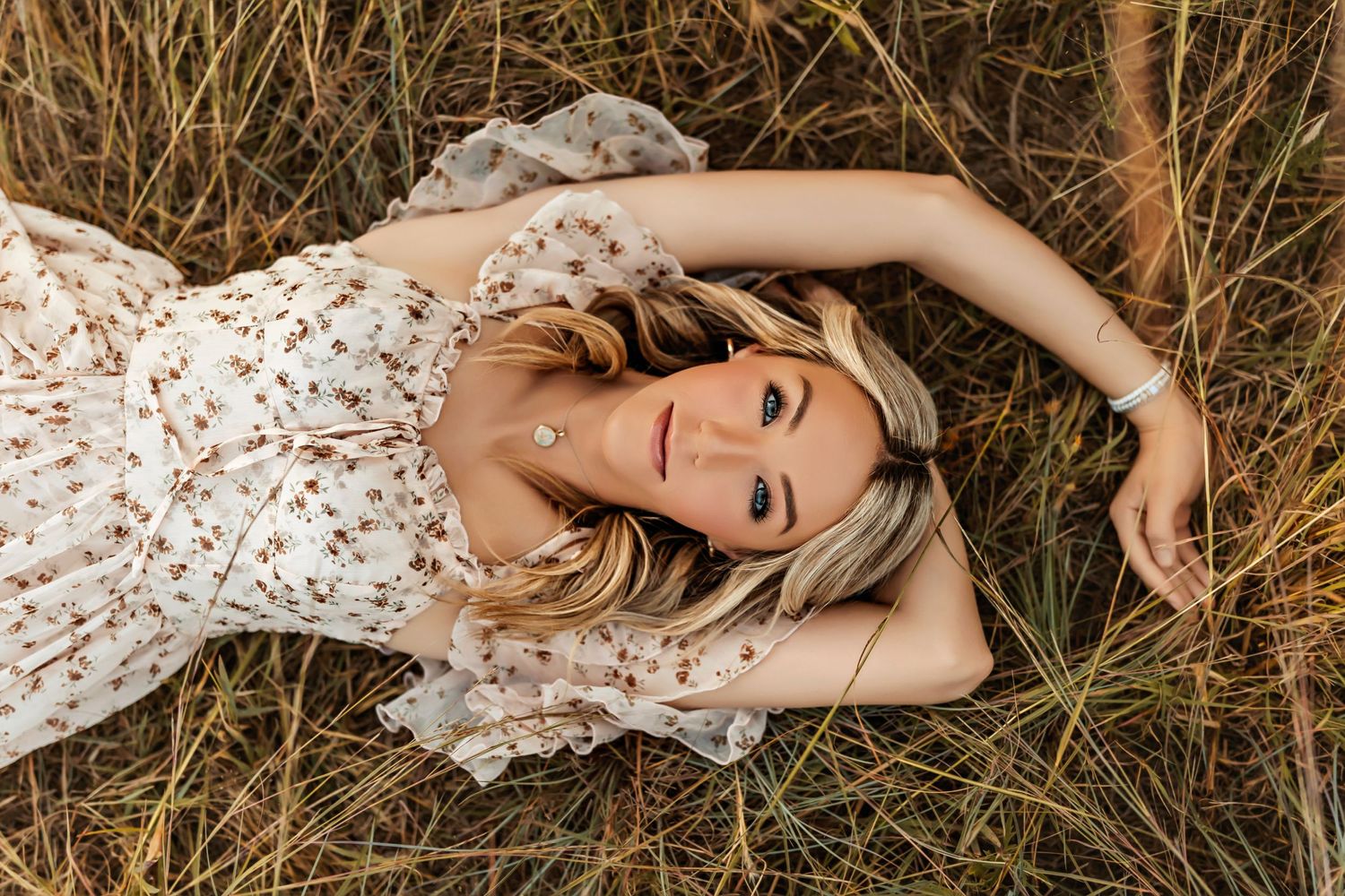 Woman in white floral dress lying in golden grass field during sunset photoshoot.