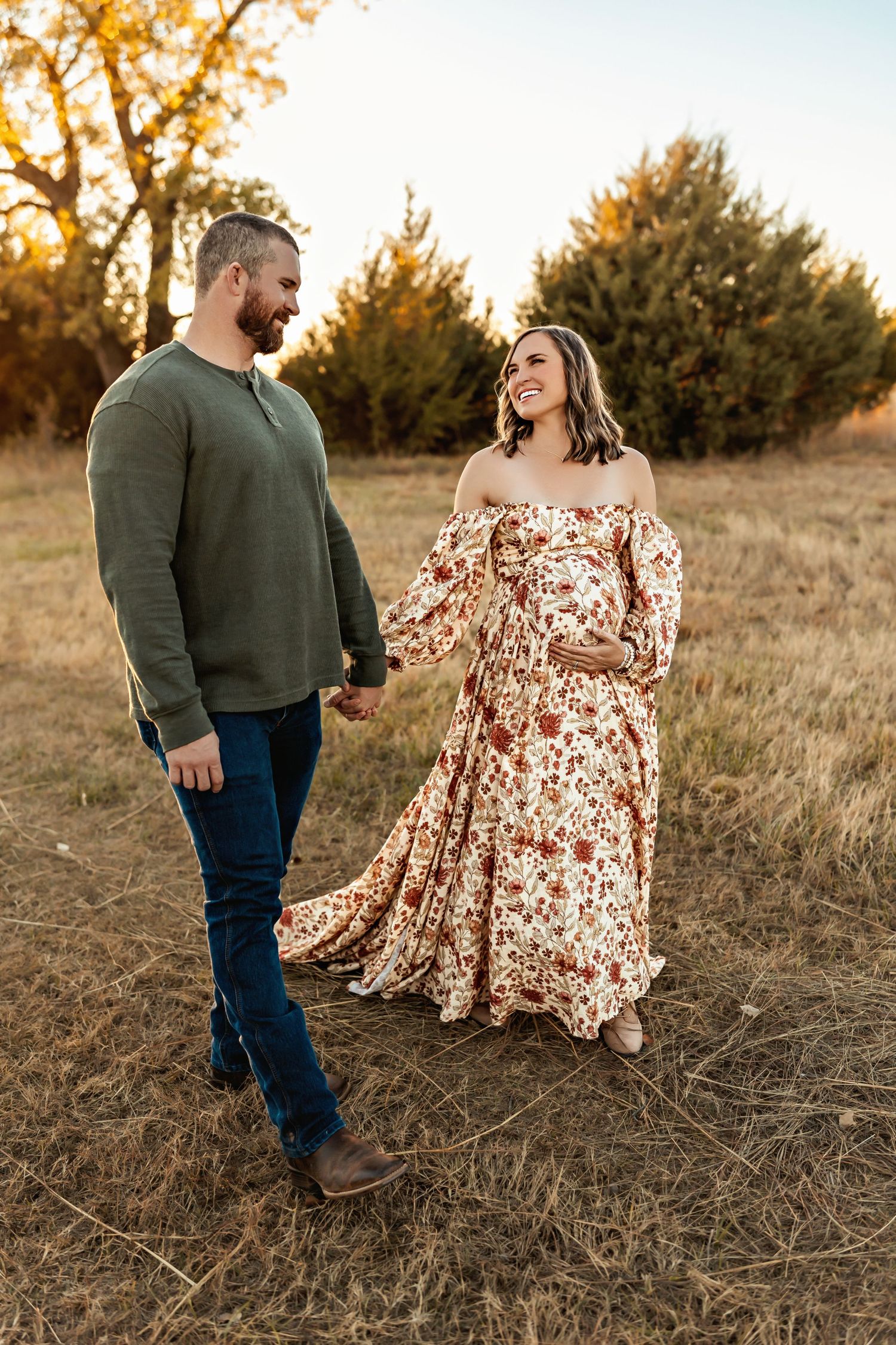 A couple stands together in a golden autumn field during sunset as her floral dress flows in the breeze.