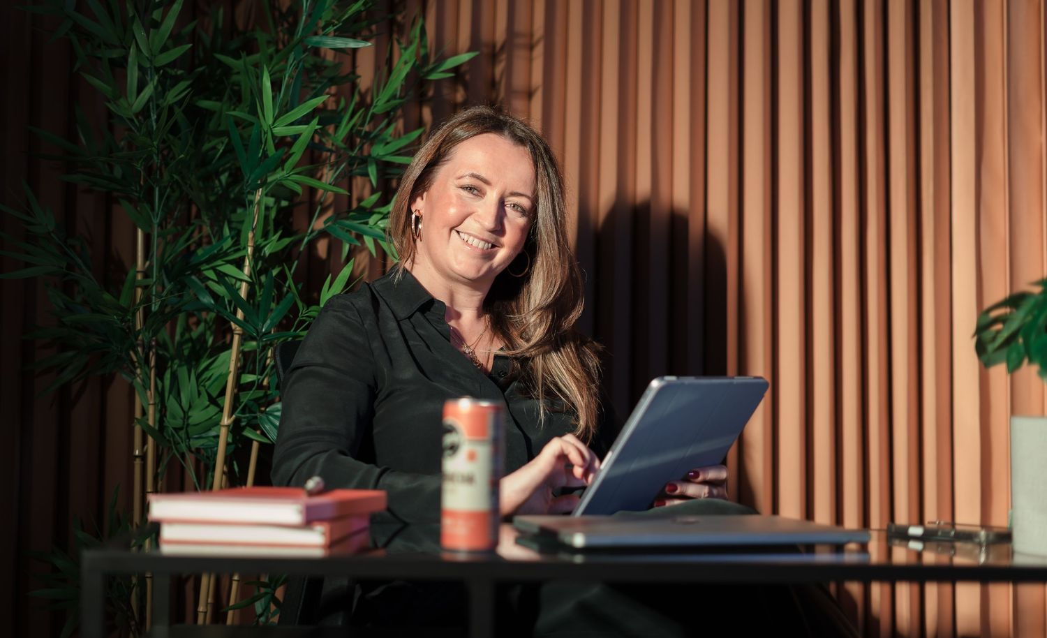 Smiling woman working on a laptop at a glass desk with books and a drink can, bamboo plants in background.