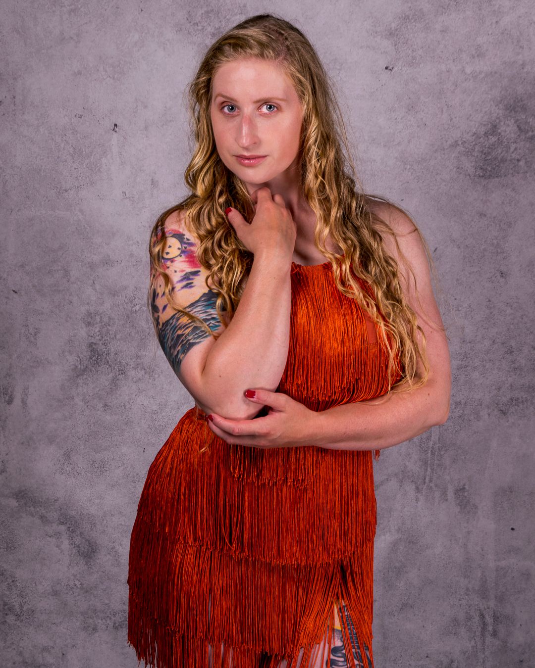 Woman in orange fringe dress with tattoos poses confidently against gray studio backdrop.
