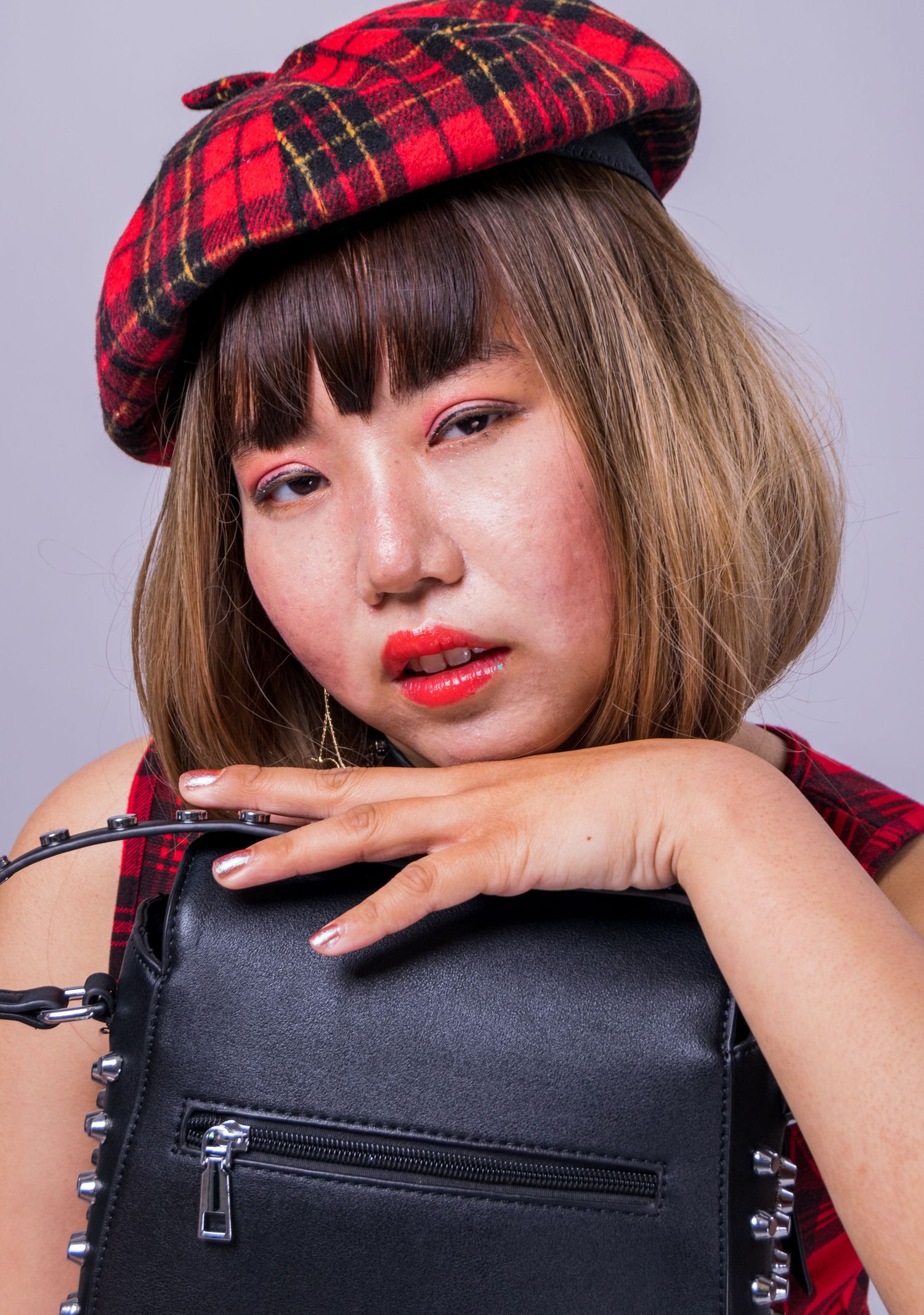 Model wearing red plaid beret poses with black handbag against gray studio background.