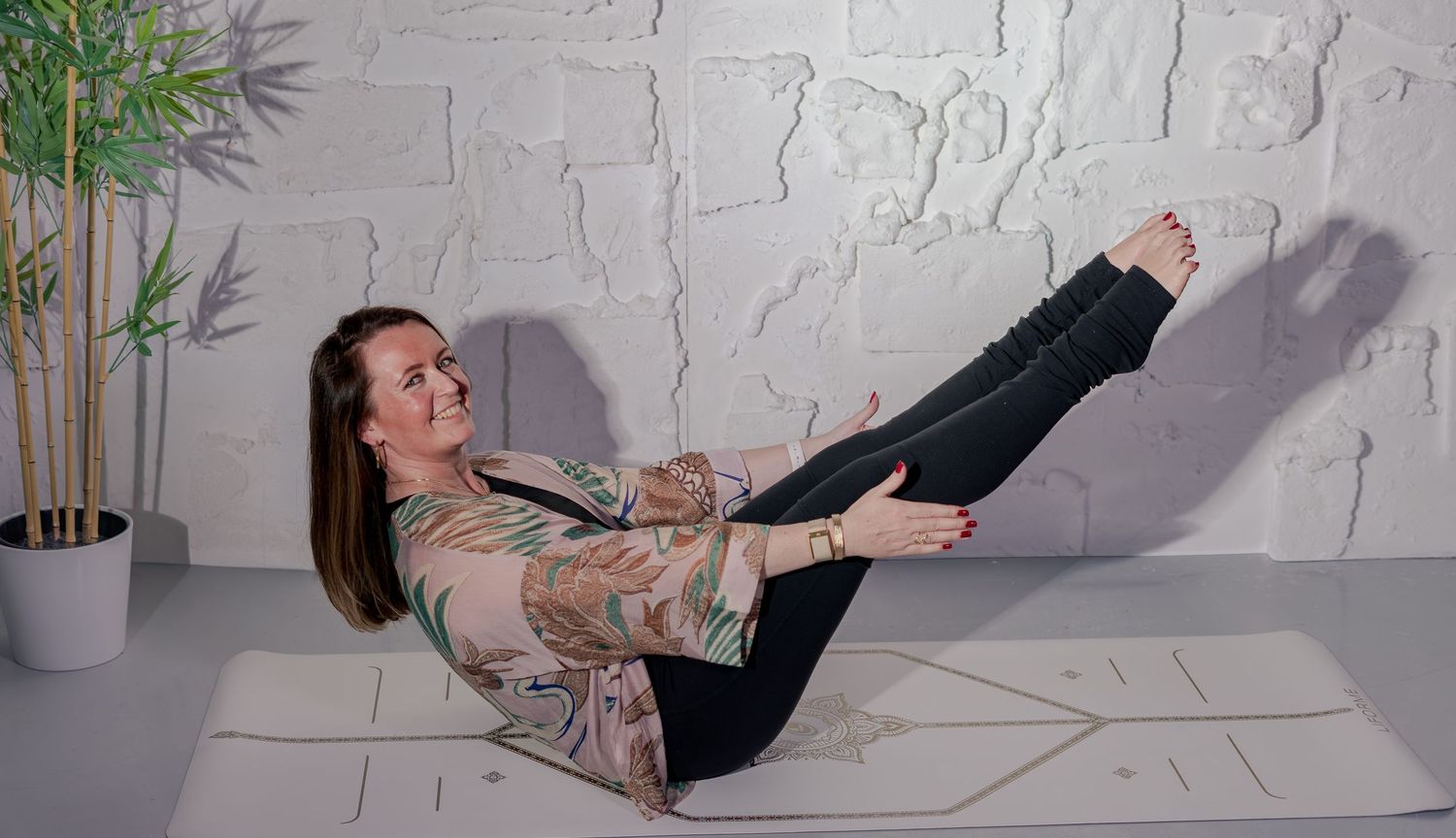 Woman practicing yoga boat pose on a mat against a white brick wall with a plant nearby.