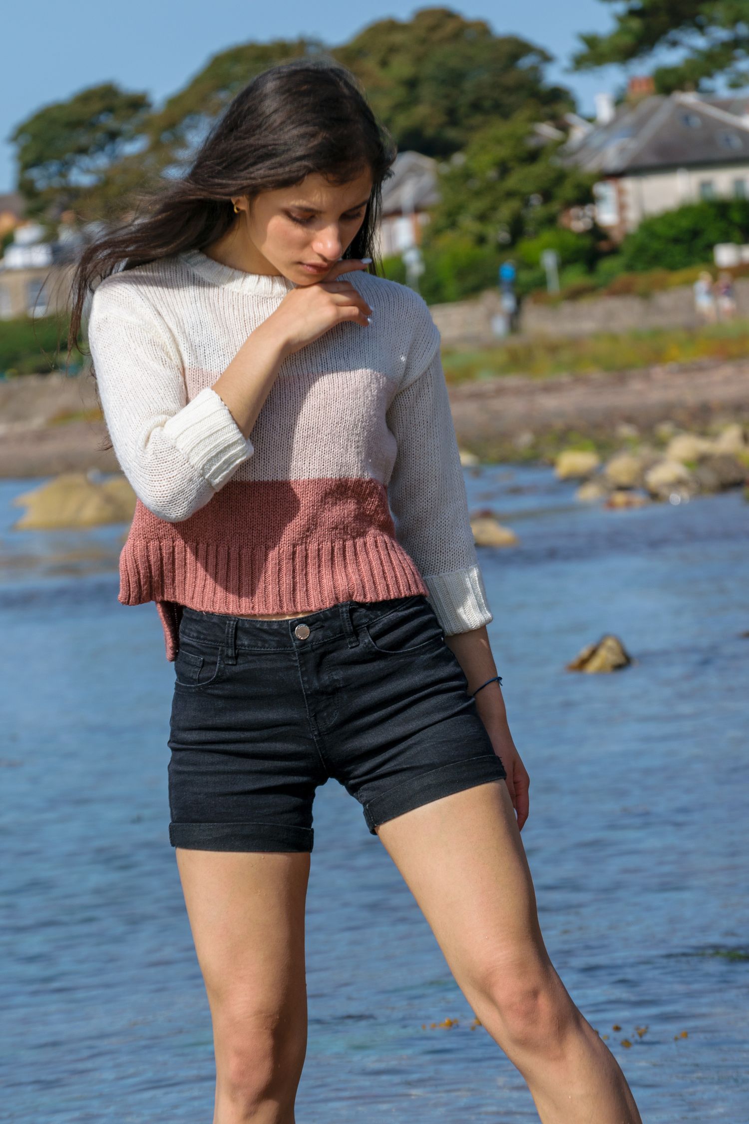 Young woman in striped knit sweater and black denim shorts standing by rocky shoreline on sunny day.