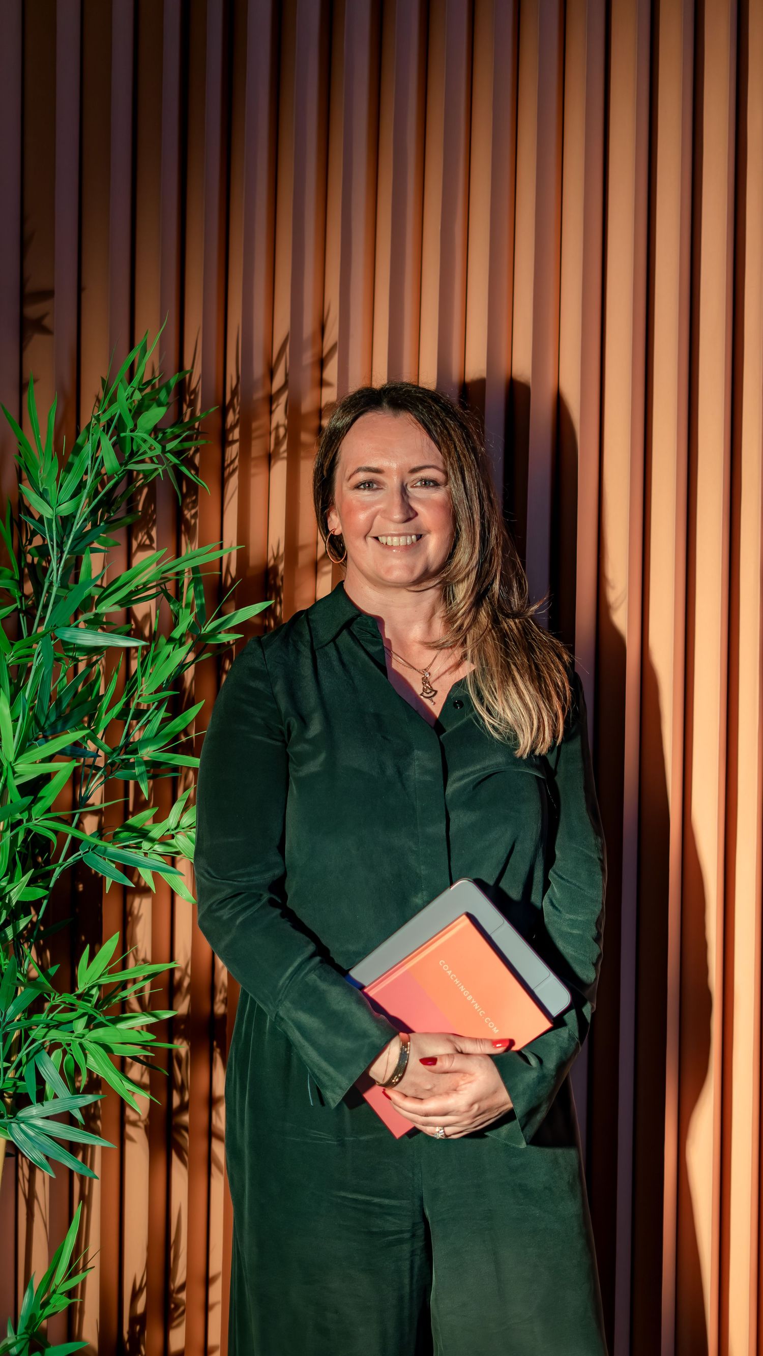 Smiling woman in green outfit holding orange books stands by wooden slat wall with bamboo plant.