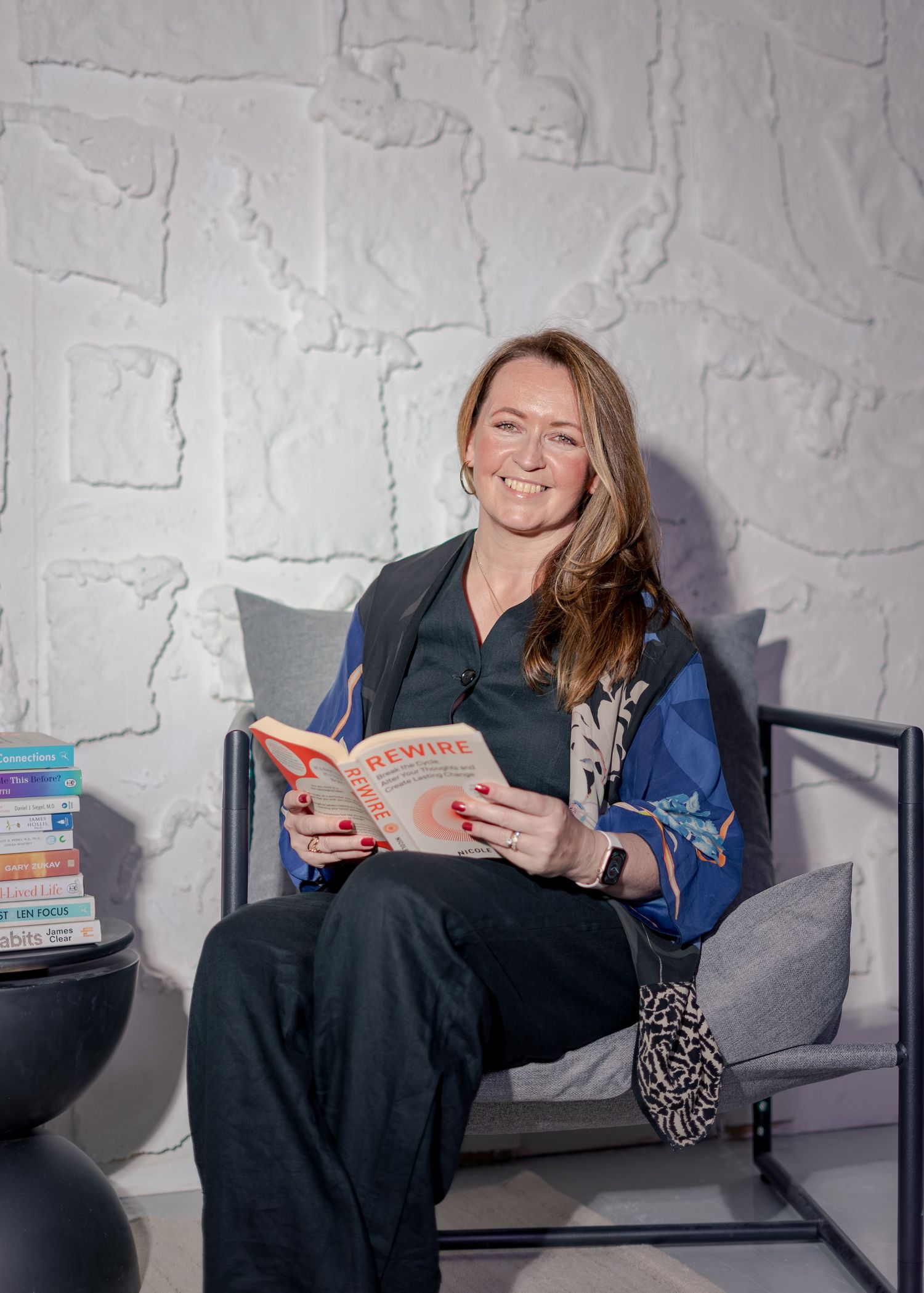 Woman smiling while reading a book titled Rewire, seated in a modern chair against a textured white wall.