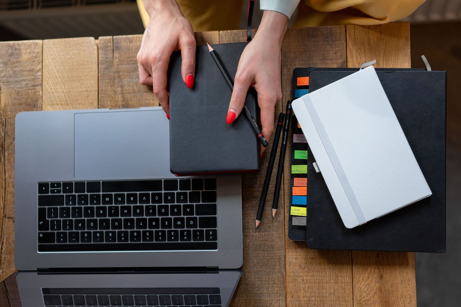A woman with red nails uses a graphics tablet beside a laptop, colorful pencils, and notebooks on a wooden desk.