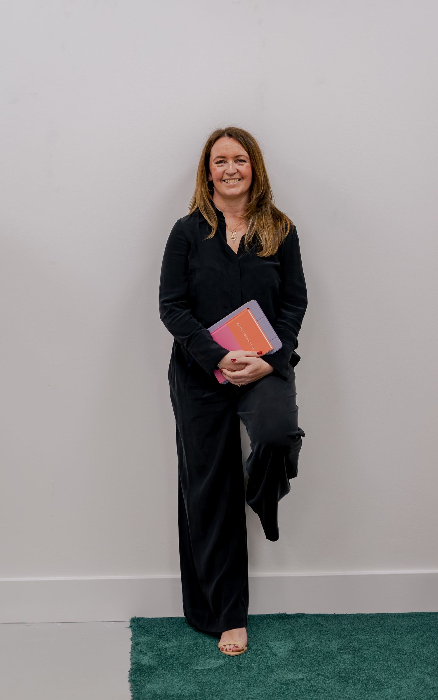 Woman in black outfit holding colorful notebook, smiling and leaning against a white wall on a teal rug.