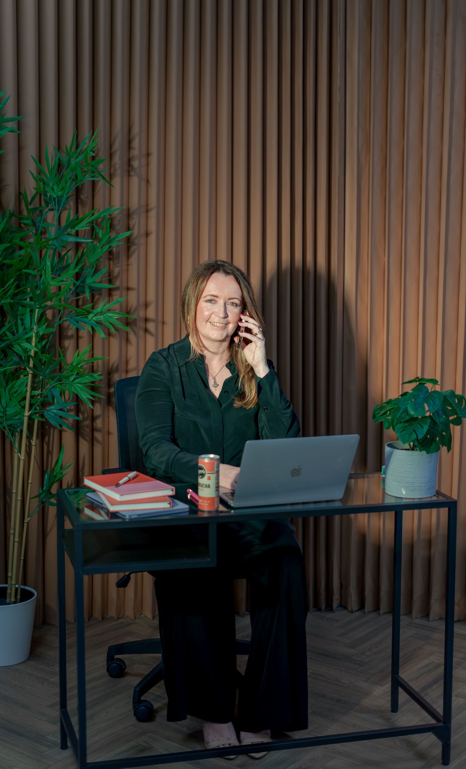 Woman in dark green suit sits at desk with laptop, talking on phone, flanked by plants in modern office.