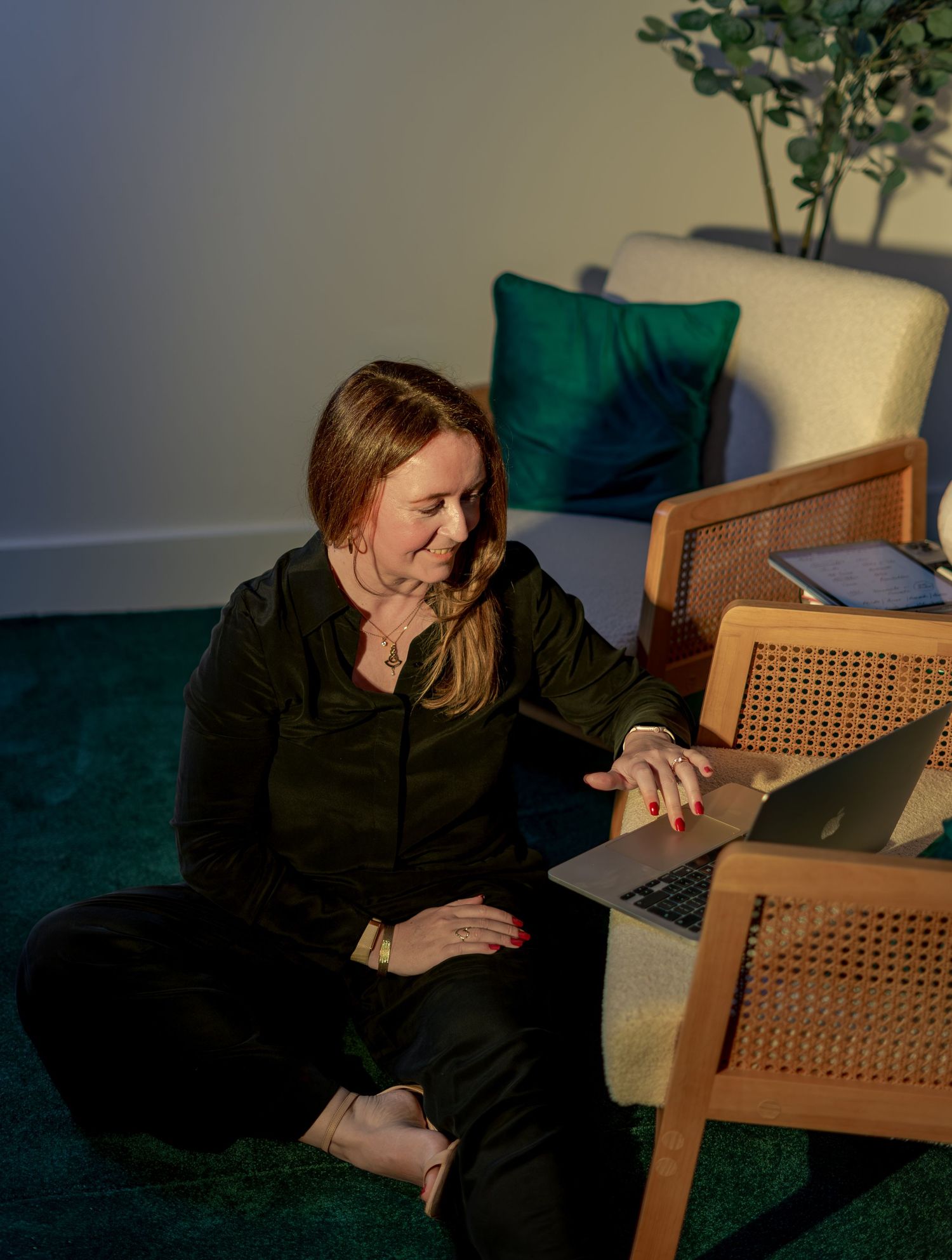 Woman in black outfit sits on green carpet, working on laptop beside a rattan chair in a cozy room.