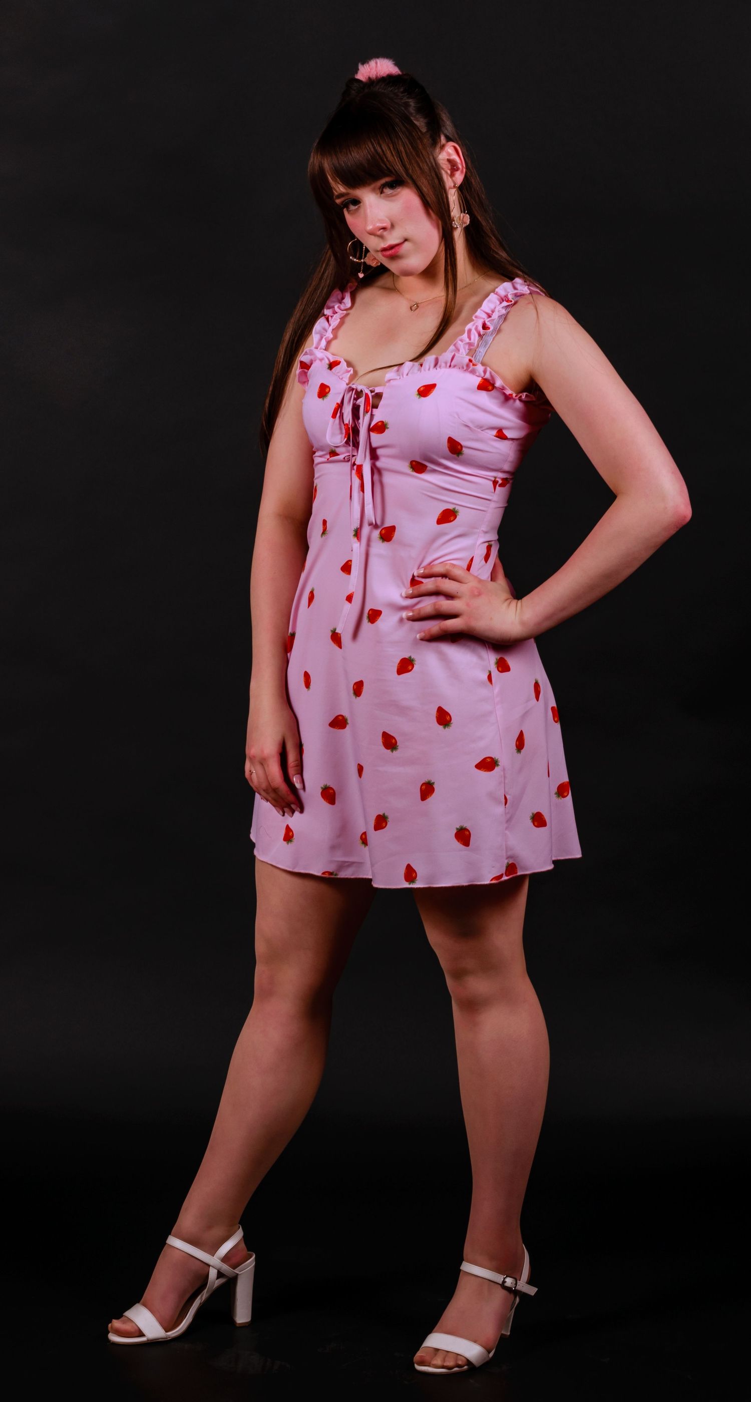 Model in pink strawberry print dress poses with white heels against black background.
