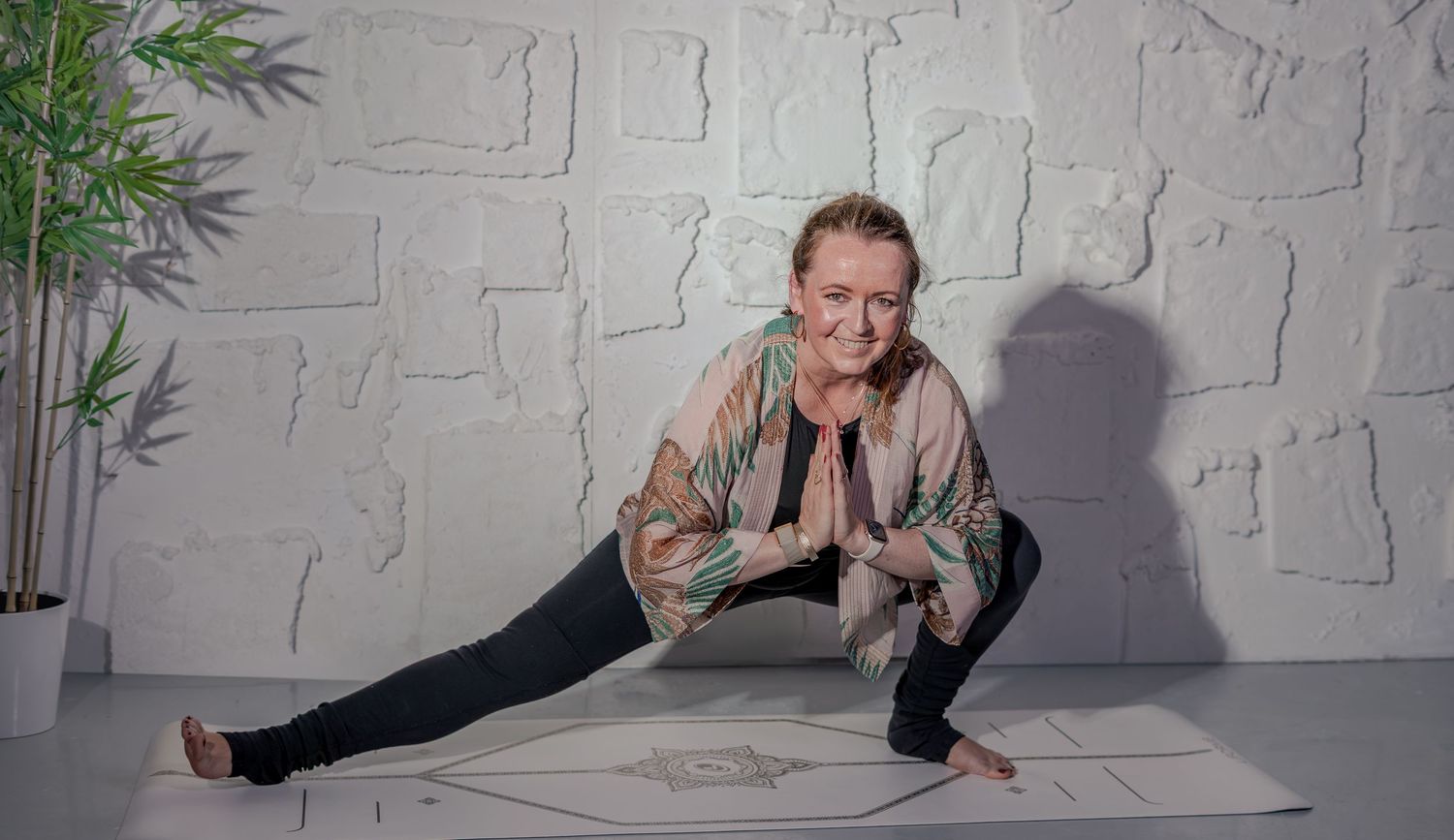Woman practicing yoga in a side lunge pose with hands in prayer position on a mat in a white studio.