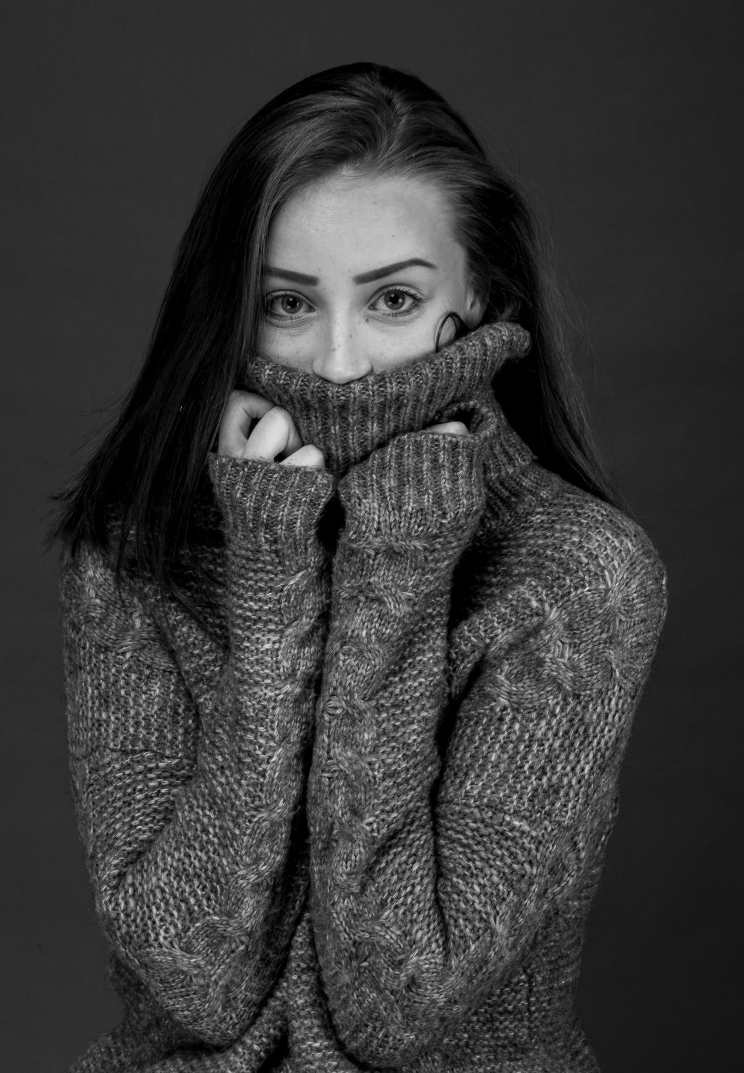 Black and white portrait of a young woman pulling a cozy knit turtleneck sweater up over her face.