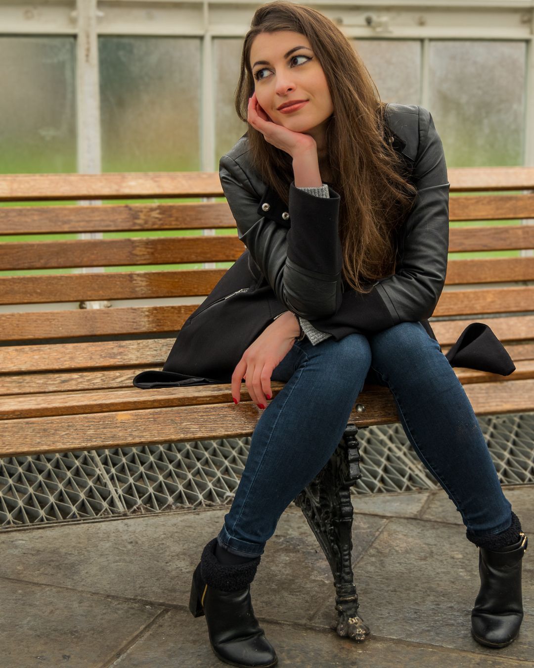 Young woman in black leather jacket and jeans sitting thoughtfully on a wooden bench wearing black boots.