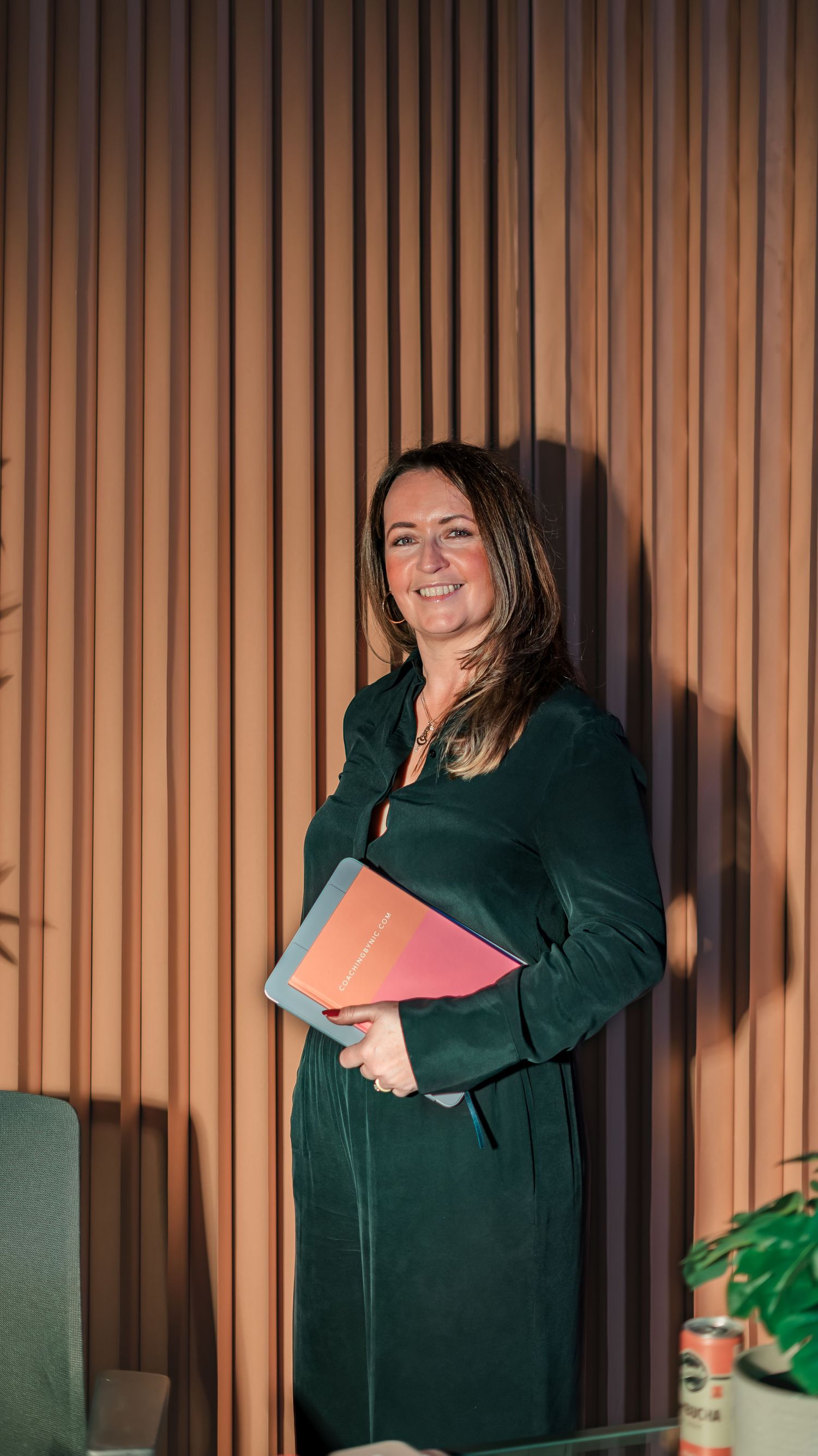 Smiling woman in green dress holding colorful folder, standing against wooden slat wall in modern office.