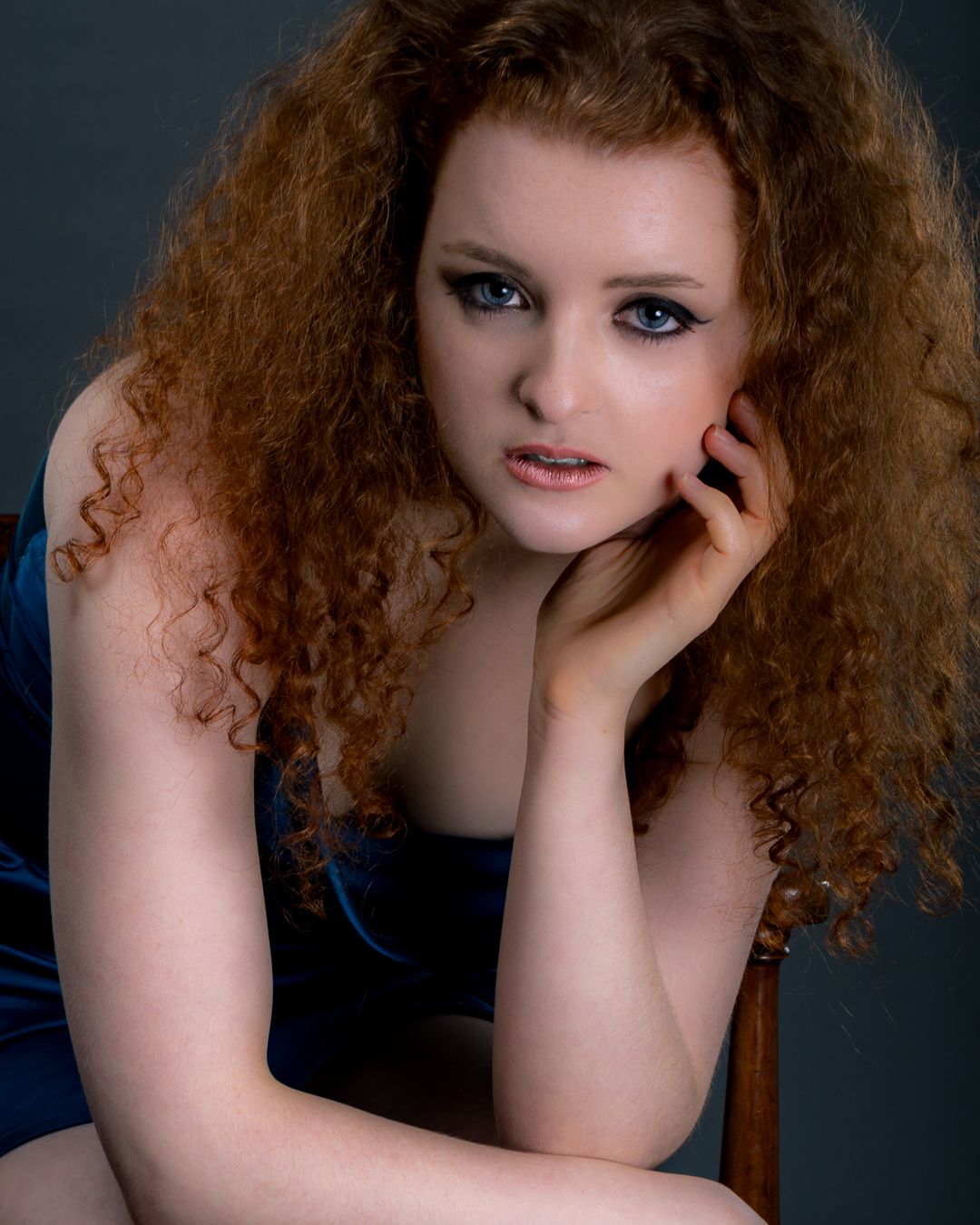 A young woman with voluminous curly red hair poses in a blue dress, resting her chin on her hand.