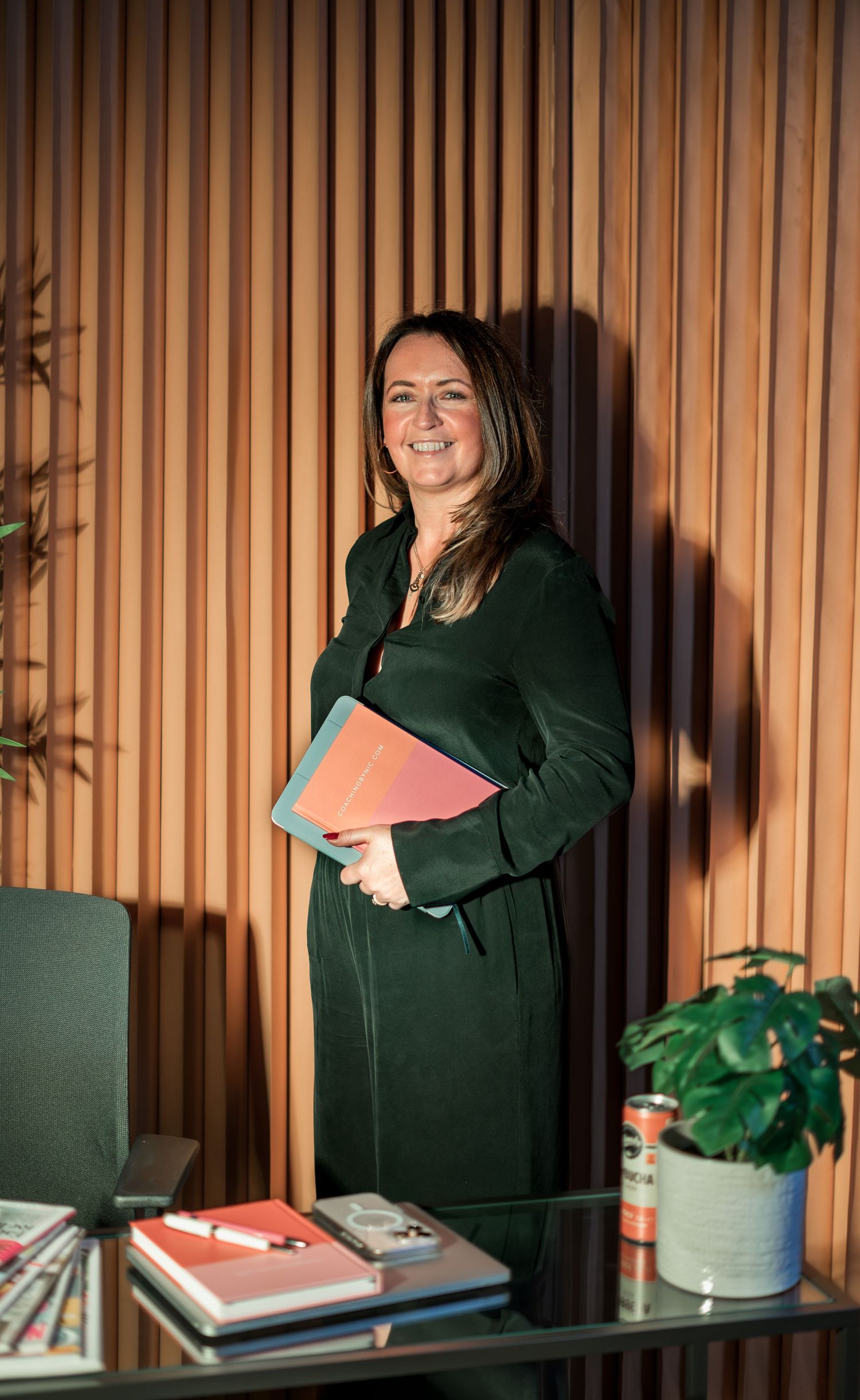 Professional woman in green dress holding books, smiling in a modern office with wooden slat wall.