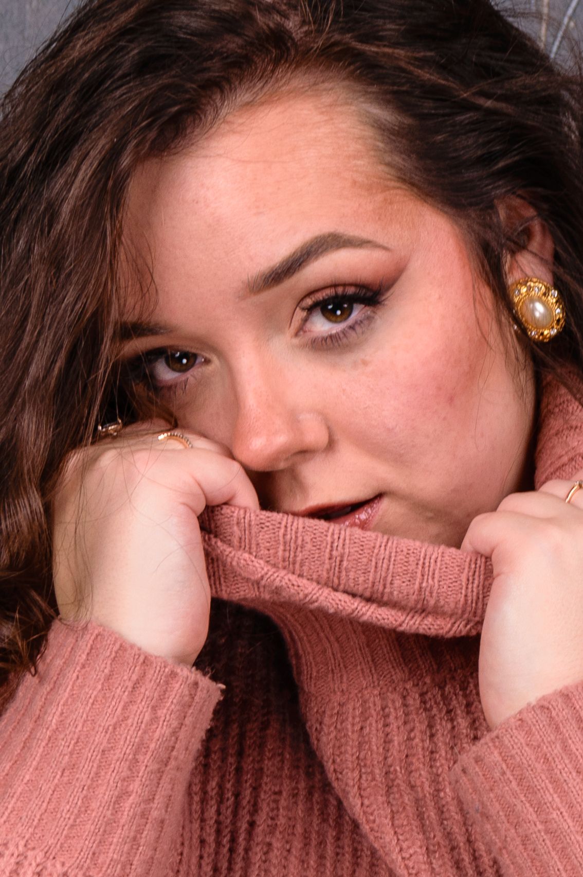 Young woman in pink turtleneck sweater pulling collar up to face, wearing gold pearl earrings and rings.