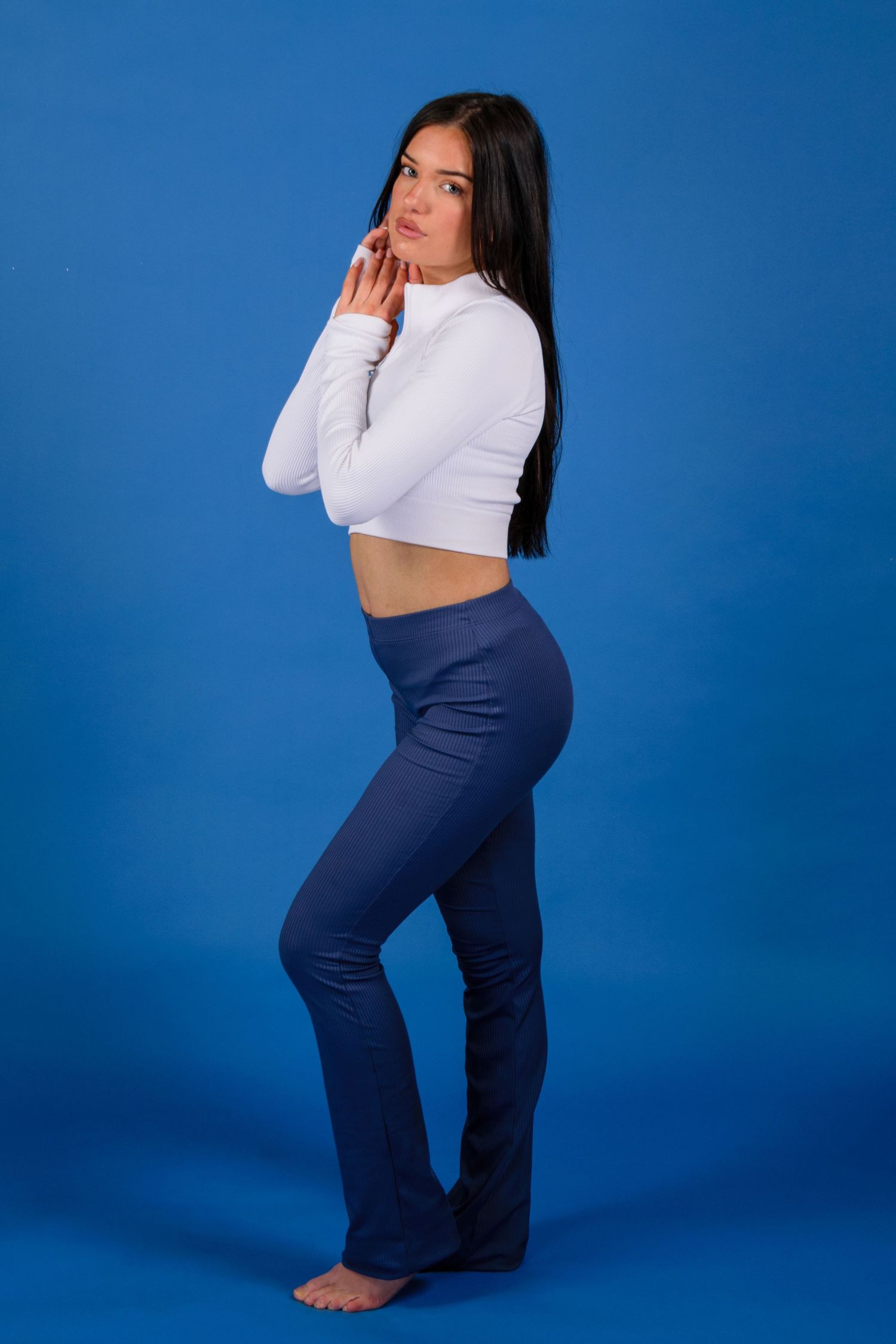 Young woman in white crop top and navy flare pants posing barefoot against a blue studio background.