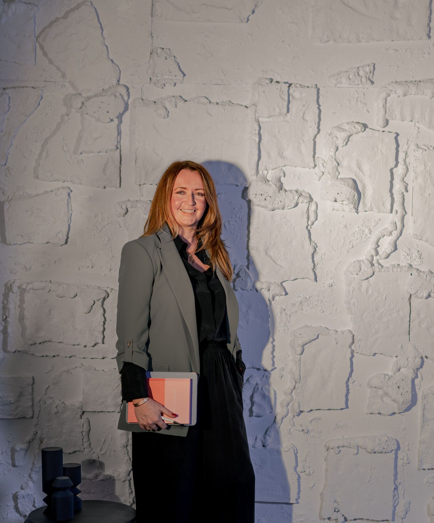 Woman in green blazer and black outfit smiling against white textured stone wall, holding a notebook.