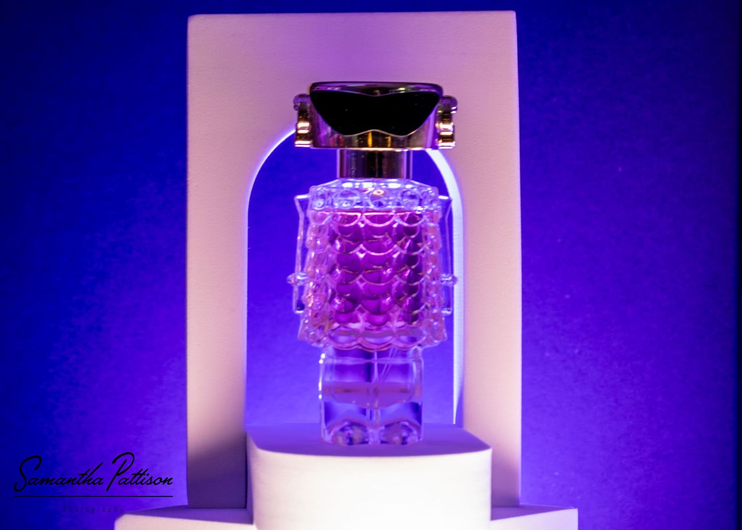 Luxury perfume bottle with pink crystal details displayed on white pedestal against vibrant purple background.