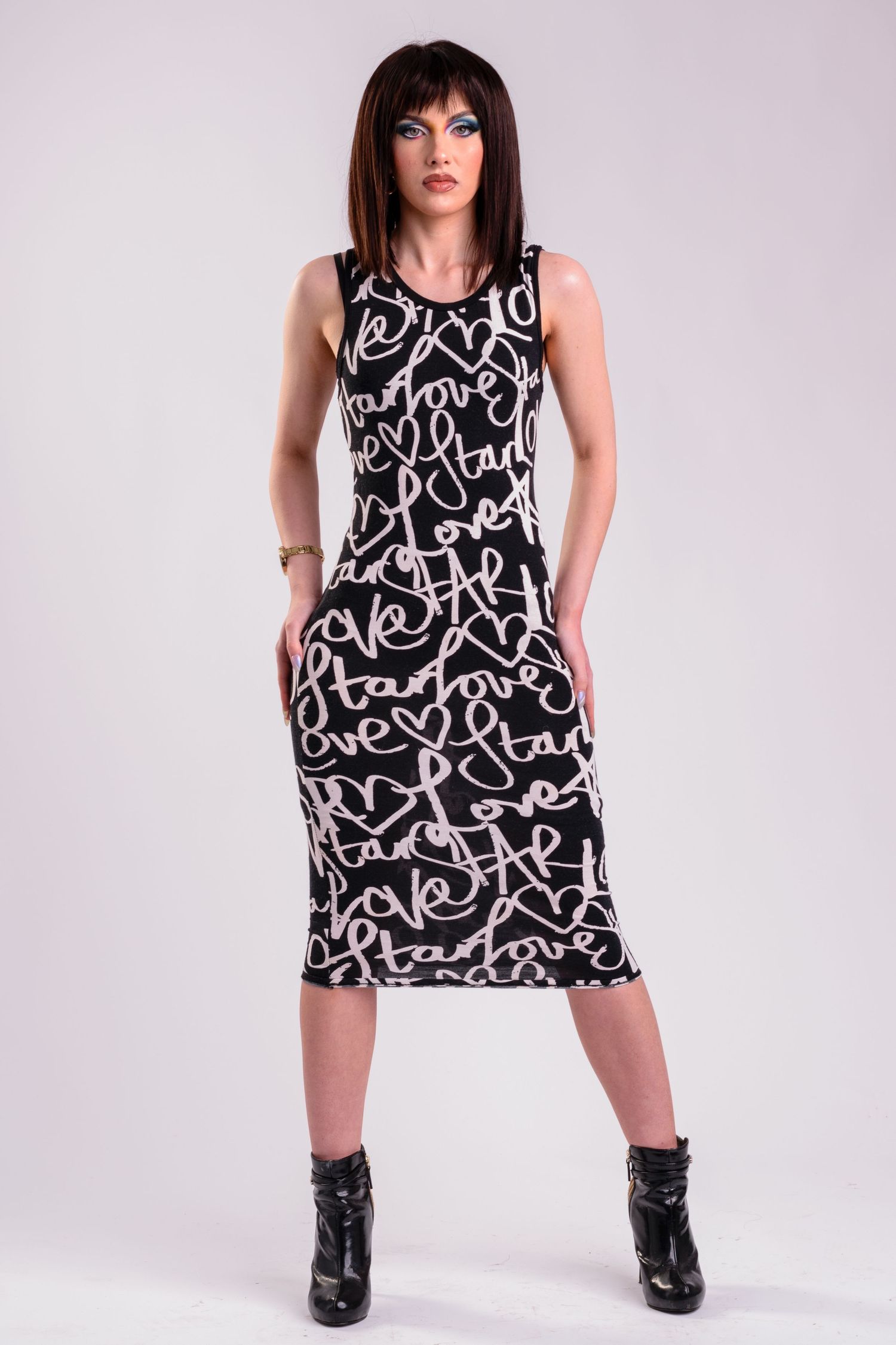 Woman wearing black midi dress with cursive love script pattern, paired with black ankle boots, posing against white background.