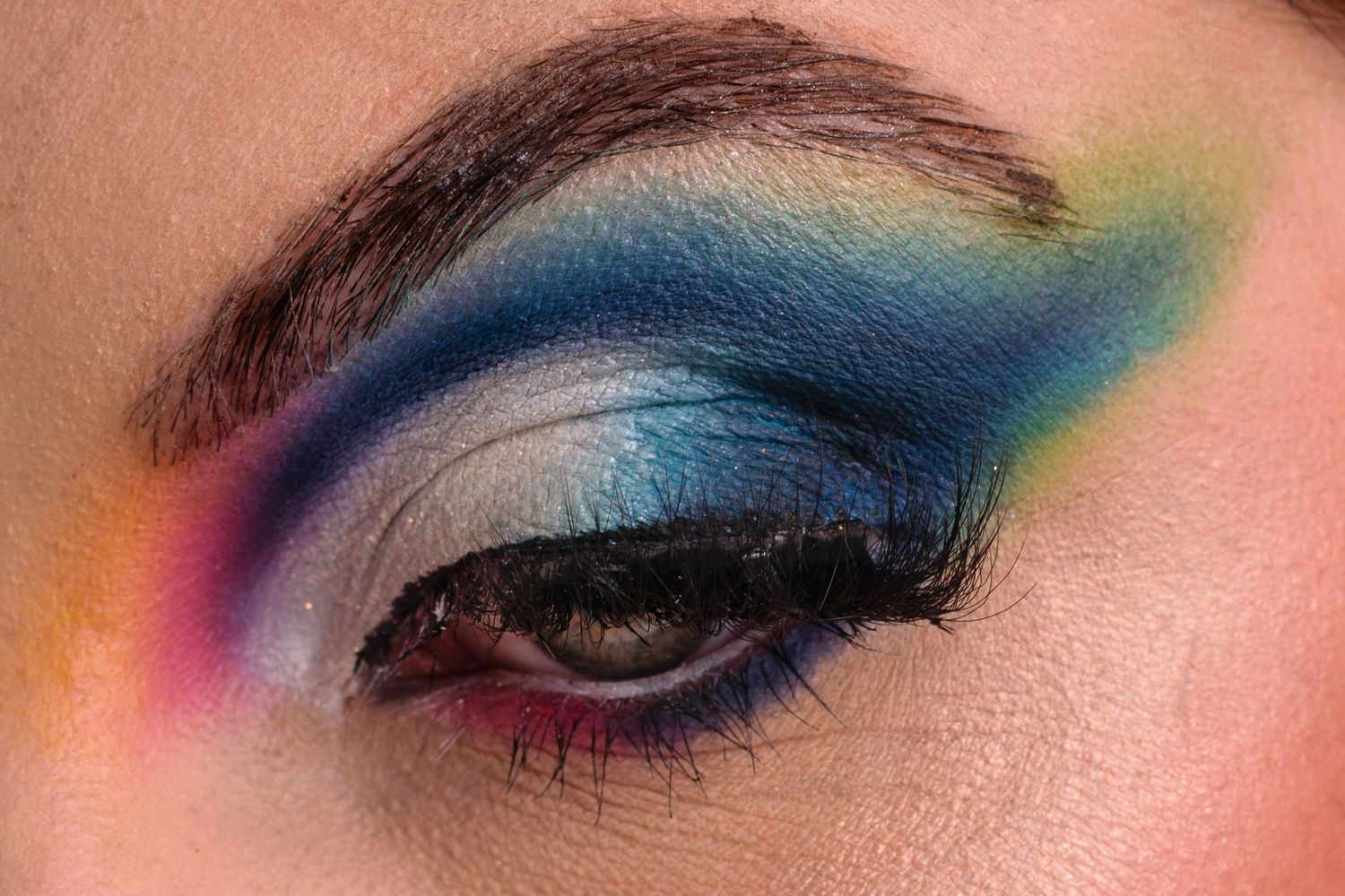 Close-up of a dramatic rainbow eye makeup look featuring blue, yellow, and pink eyeshadow with bold liner.