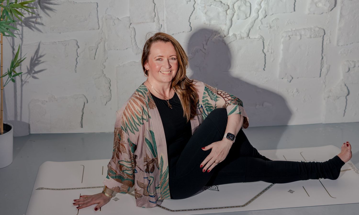 Woman in colorful kimono jacket sits relaxed on yoga mat in bright white studio, smiling warmly.