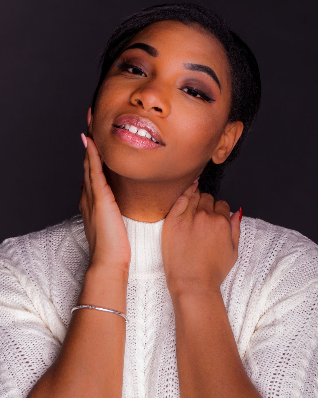 Young Black woman in white sweater poses gracefully, hands near face, looking up against dark background.